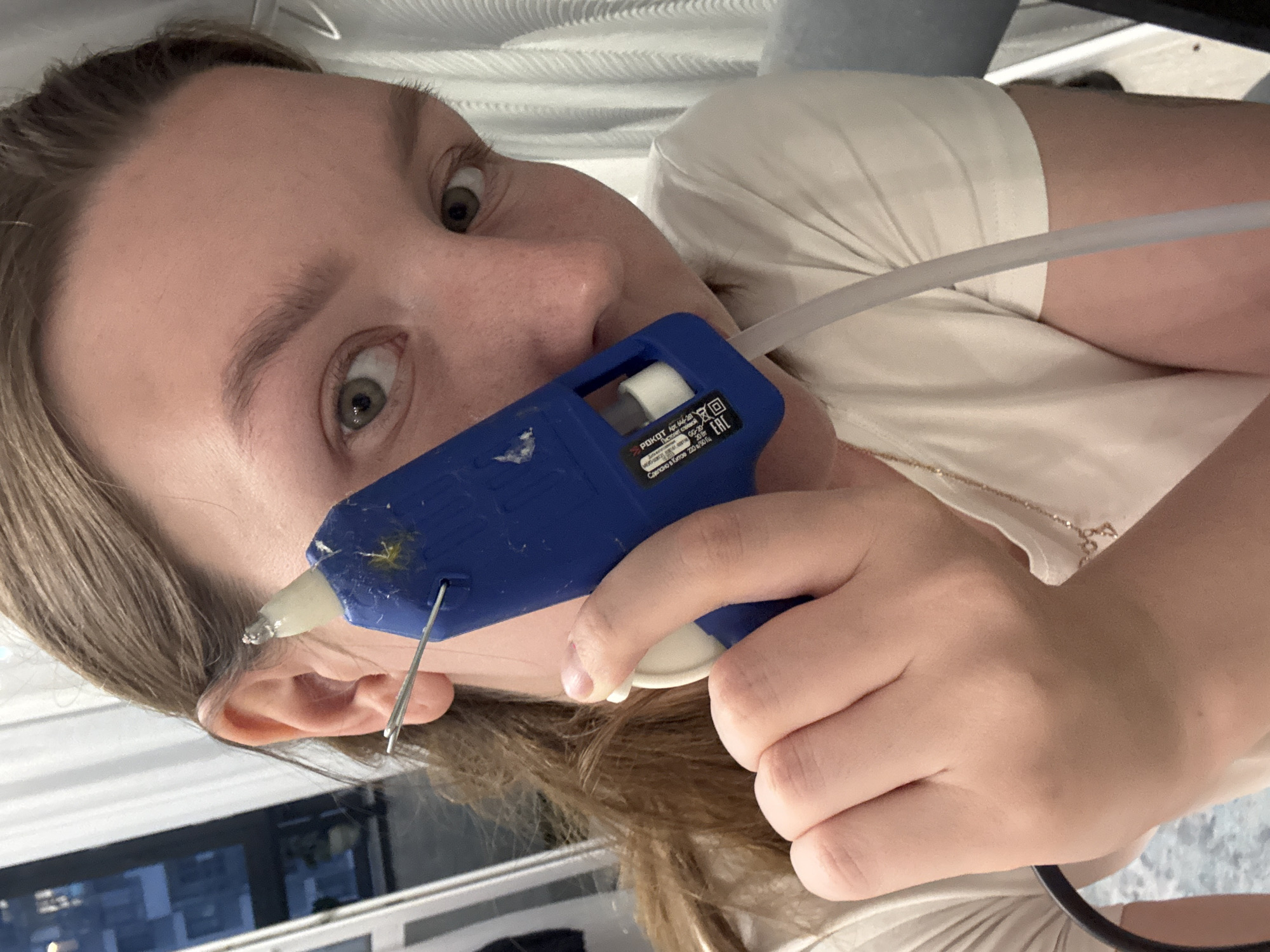 The image shows a close-up of a young woman's face and hand, holding a blue hot glue gun near her ear. She has light brown hair and green eyes. A metal wire protrudes from her ear, with the hot glue gun nozzle positioned very close to it. Her left hand is visible, holding the glue gun. She is wearing a white t-shirt with a delicate gold necklace. The background is blurry but appears to be an indoor setting with curtains and possibly a window showing a city view with buildings, which could be Izhevsk, Russia. The overall lighting suggests it is nighttime or dusk.