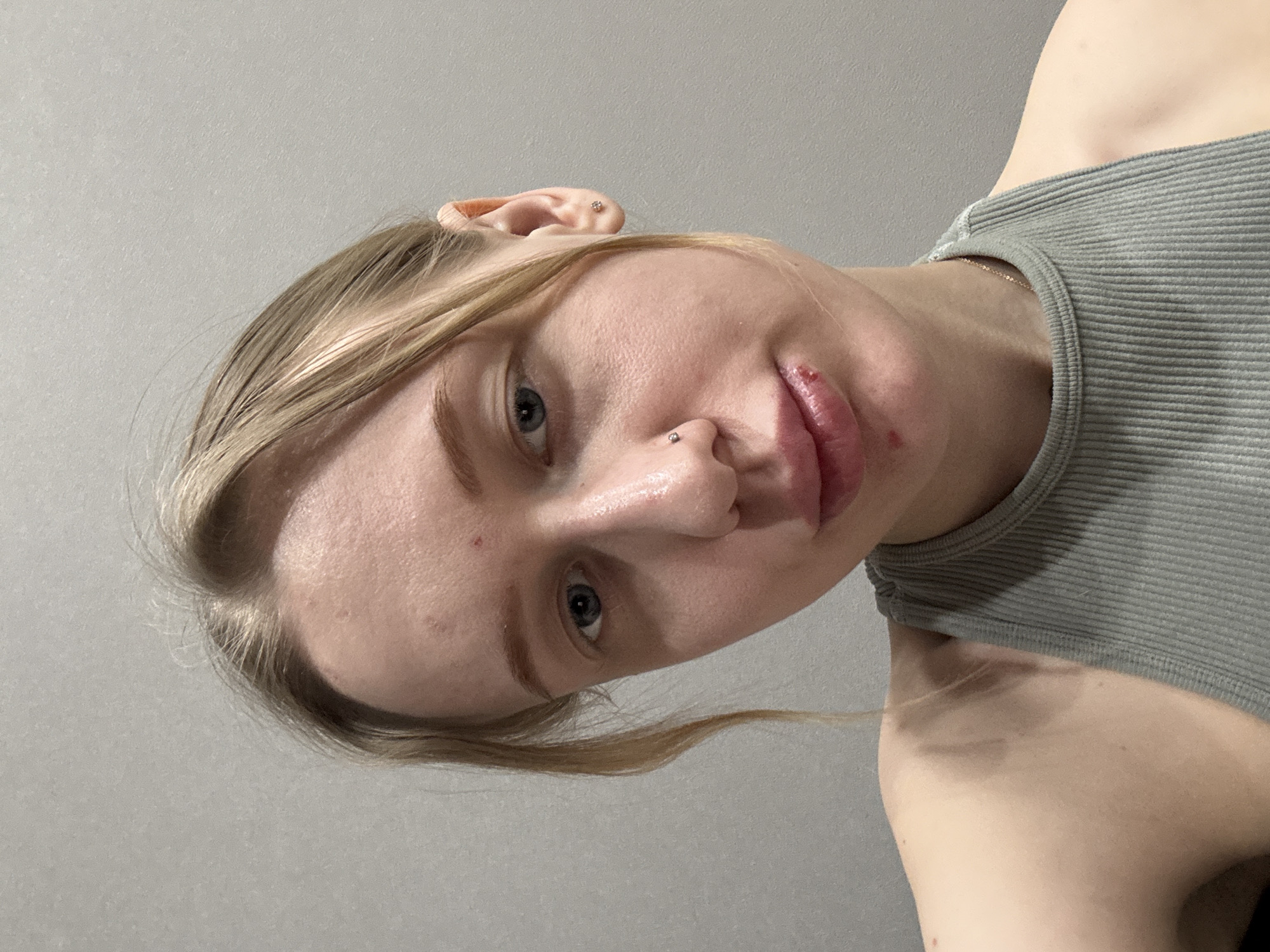 A close-up, angled shot of a fair-skinned person with light blonde hair pulled back, facing the camera. The person has light blue eyes, a nose piercing, and appears to have chapped lips. A small blemish is visible on their cheek. They are wearing a grey ribbed tank top. The background is a plain grey wall. The location in the city of Rostov-on-Don, Russia is indeterminate from the image.
