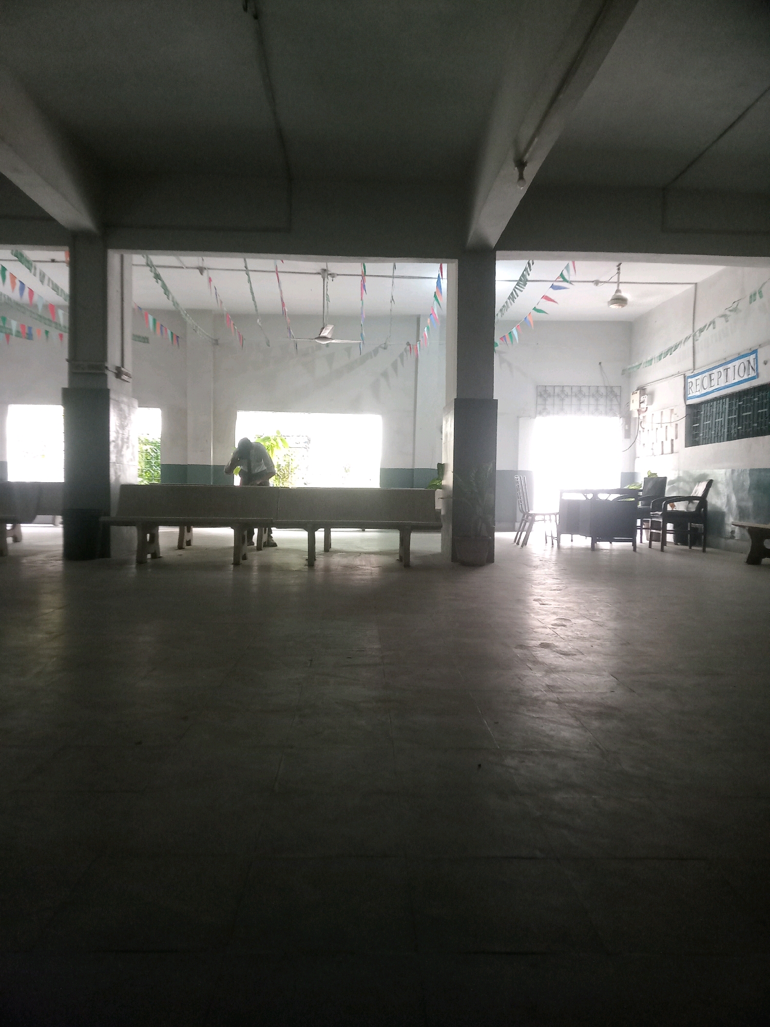 The image depicts a long, indoor corridor or hallway, likely within an institutional building such as a school or office. The space is sparsely furnished with concrete benches running along the walls and a reception desk area with chairs visible in the background. Festive buntings are strung across the ceiling, suggesting a celebratory event or decoration. A person is seen standing and interacting with something on one of the benches in the mid-ground. The lighting is dim, with sunlight filtering in from windows at the far end of the hallway, creating shadows on the concrete floor. The overall impression is one of a functional, yet decorated, common area. The location is the city of Karachi Division, Pakistan.