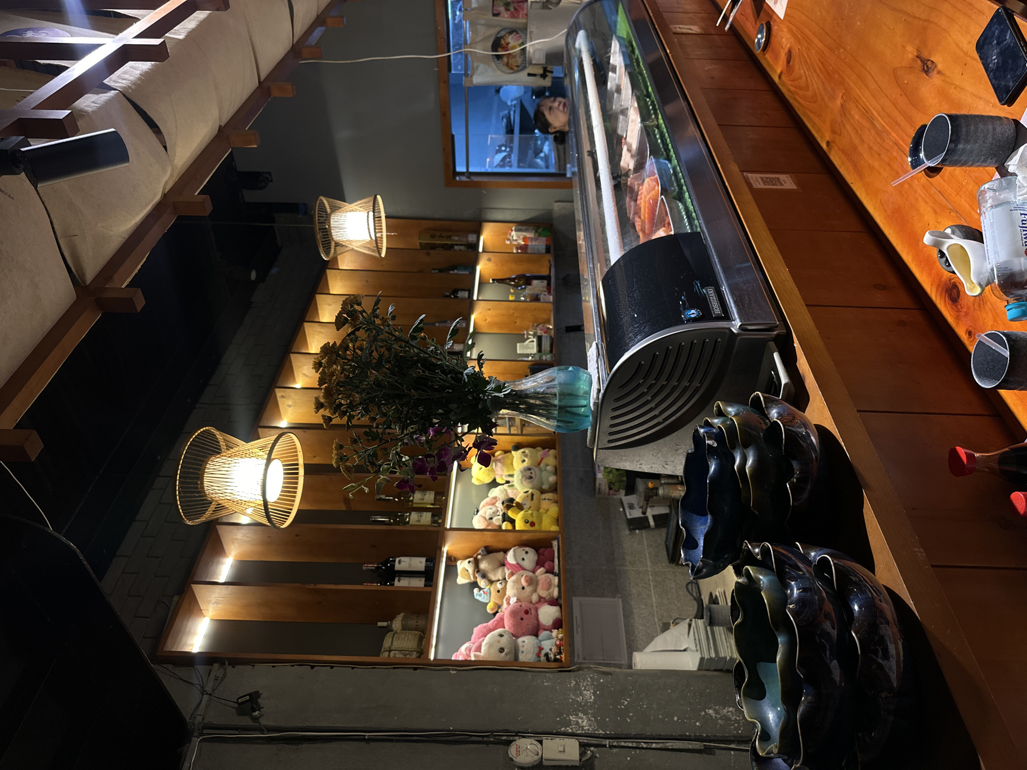 The image depicts the interior of a cozy restaurant, likely a sushi or Japanese eatery, with a warm and intimate ambiance. The primary perspective is from a diner's seat, looking across a counter and towards a wall decorated with shelves.
To the right, a wooden counter extends, featuring several practical items. Black cylindrical holders, possibly for chopsticks or straws, are present, one holding a clear plastic bottle with a blue cap and a straw. A small white and yellow ceramic jug, likely for soy sauce, is also on the counter. A large, rectangular refrigerated display case, bearing the "HOSHIZAKI" brand name, occupies a significant portion of the counter, showcasing various cuts of raw fish or sashimi, indicating the establishment's cuisine. Stacked dark, wavy ceramic bowls are also visible near the edge of the counter.
To the left, a multi-tiered wooden shelving unit extends upwards, creatively lit from within by strip lights. This unit holds various bottles, possibly of wine or sake, and other decorative items. Notably, two of the larger compartments are filled with an assortment of plush toys, including teddy bears, a Pikachu, and other animal figures, adding a whimsical touch to the decor. On top of these shelves, a vase holds artificial flowers, a mix of purple and dried-looking yellow blooms. Two distinct hanging light fixtures with intricate, woven wooden shades emit a warm, golden glow, contributing significantly to the inviting atmosphere.
In the background, through an opening or window that appears to lead into a kitchen area, a person's head is visible, looking out into the dining space. Above the shelving unit, the ceiling features a sloped design with wooden beams and light-colored fabric or canvas panels, illuminated by hidden or recessed lighting. The overall lighting is dim, suggesting it is evening or night, enhancing the relaxed and pleasant mood of the restaurant.