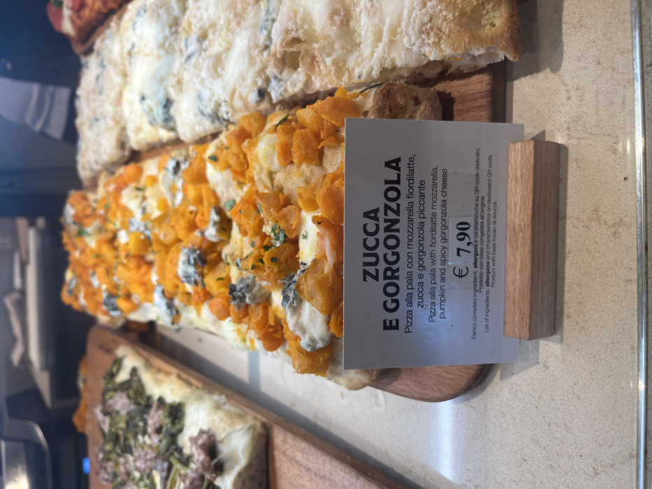 This media file captures a close-up view of various slices of pizza alla pala displayed in what appears to be a food counter, likely a pizzeria or bakery, in Dorno, Italy. The main subject is a rectangular slice of pizza topped generously with diced orange pumpkin (zucca), chunks of blue-veined gorgonzola cheese, and finely chopped green herbs, all resting on a creamy white base and focaccia-style dough.
Behind this prominent slice, another pizza with a lighter, perhaps cheesy or herb-based topping is partially visible. Further in the background, another pizza slice with darker, possibly vegetable or meat toppings, can be discerned, though it is out of focus. All pizzas are presented on light-colored wooden cutting boards, emphasizing a rustic and artisanal aesthetic. The setting suggests an indoor commercial environment with bright, possibly artificial, lighting illuminating the food.
A white price tag is strategically placed next to the pumpkin and gorgonzola pizza. The visible text on the tag reads:
* "ZUCCA E GORGONZOLA"
* "Pizza alla pala con mozzarella fiordilatte, zucca e gorgonzola piccante"
* "Pizza alla pala with fiordilatte mozzarella, pumpkin and spicy gorgonzola cheese"
* "€ 7,90"
* Smaller text at the bottom indicates: "Elenco completo ingredienti, allergeni e caratteristiche su QR code dedicato. Prodotto con base congelata all'origine. List of ingredients, allergens and characteristics on dedicated QR code. Product with base frozen at source."
The image depicts a selection of freshly prepared pizza slices, highlighting their ingredients and pricing for customers, consistent with a food service establishment in Italy. No people are visible in the frame.