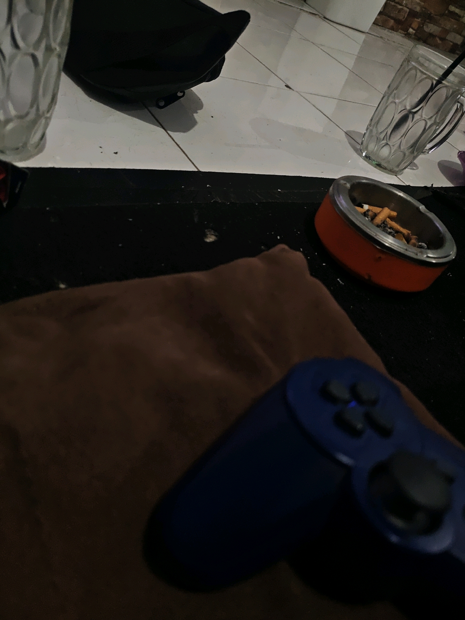 In a dimly lit indoor setting, likely a home or a casual establishment, a blue video game controller rests on a brown, textured surface, possibly a blanket or sofa. The controller is angled towards the viewer, with its buttons and analog sticks visible. In the background, an orange ashtray filled with cigarette butts sits on a black surface. To the left, a clear glass with a ribbed pattern is partially visible. The floor is tiled and appears somewhat dirty, with small dark particles scattered across it. A black bag is also visible in the upper left corner. The overall scene suggests a person is about to engage in or is in the middle of a gaming session, accompanied by smoking. The context of Pringsewu, Indonesia, implies this could be a typical domestic scene in that region, potentially in a living room or a recreational area.