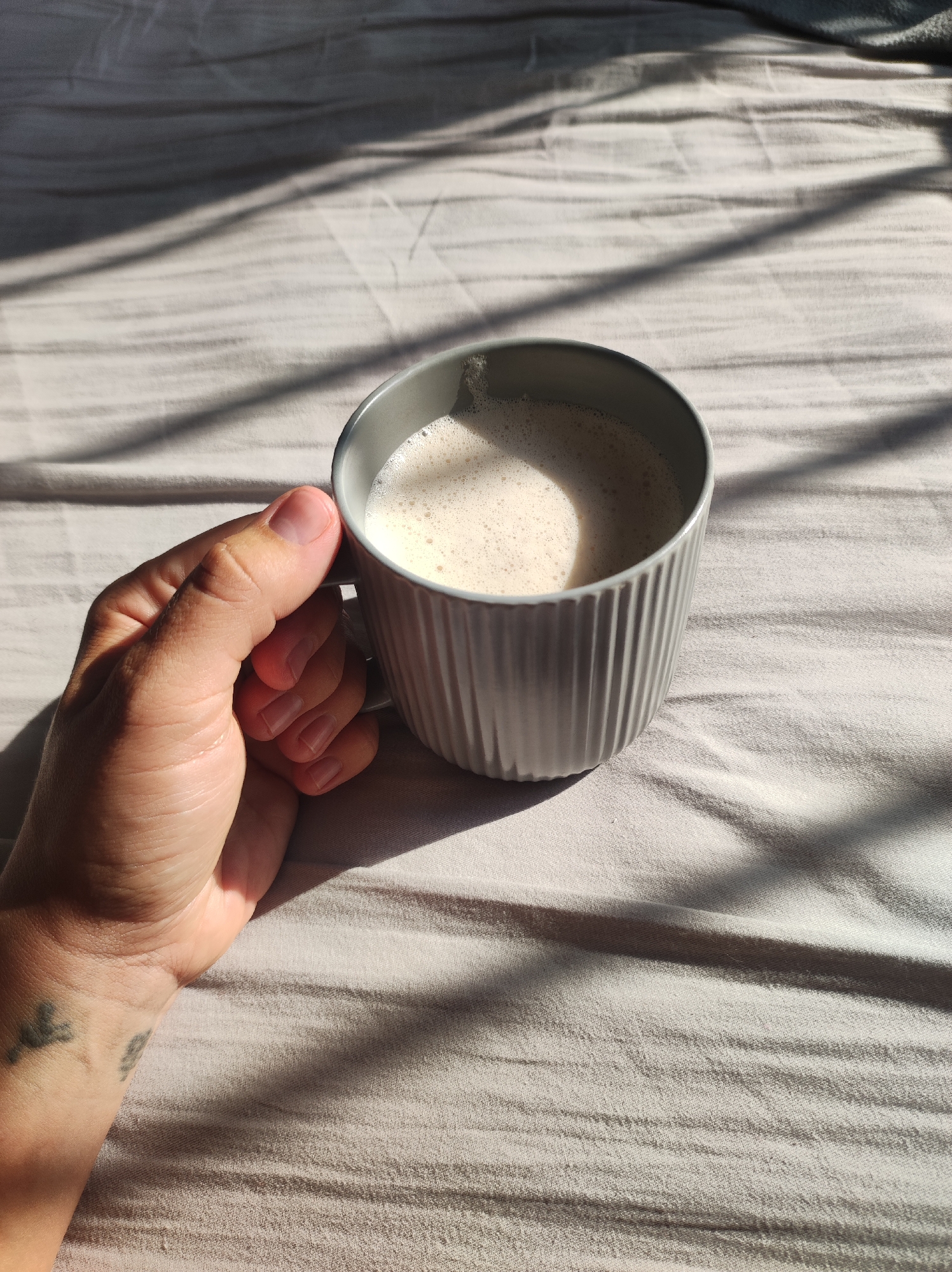 A hand holds a grey, ribbed mug filled with a light-colored, frothy beverage. The mug rests on a light-colored, textured surface, possibly bedding, which is illuminated by sunlight casting diagonal shadows across it. The scene is domestic, with no indication of a specific city or location.