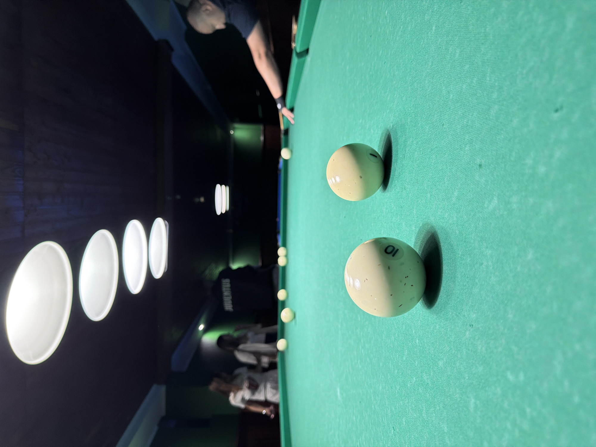 The image depicts an indoor pool hall, likely located in Strășeni, Moldova, though this cannot be definitively confirmed from the visual information alone. The scene focuses on a green felt billiard table with several white balls scattered across it. In the foreground, two billiard balls are prominently displayed, one of which is numbered '10'. To the right, a man wearing a dark t-shirt is actively playing pool, aiming a cue stick towards a ball in the distance. In the background, several blurry figures, appearing to be two women and a man wearing a "JUVENTUS" branded shirt, are visible standing behind the table. The environment is dimly lit by overhead pendant lights, creating a moody atmosphere. The overall impression is of a casual gathering or game in progress within a dedicated recreational space.
