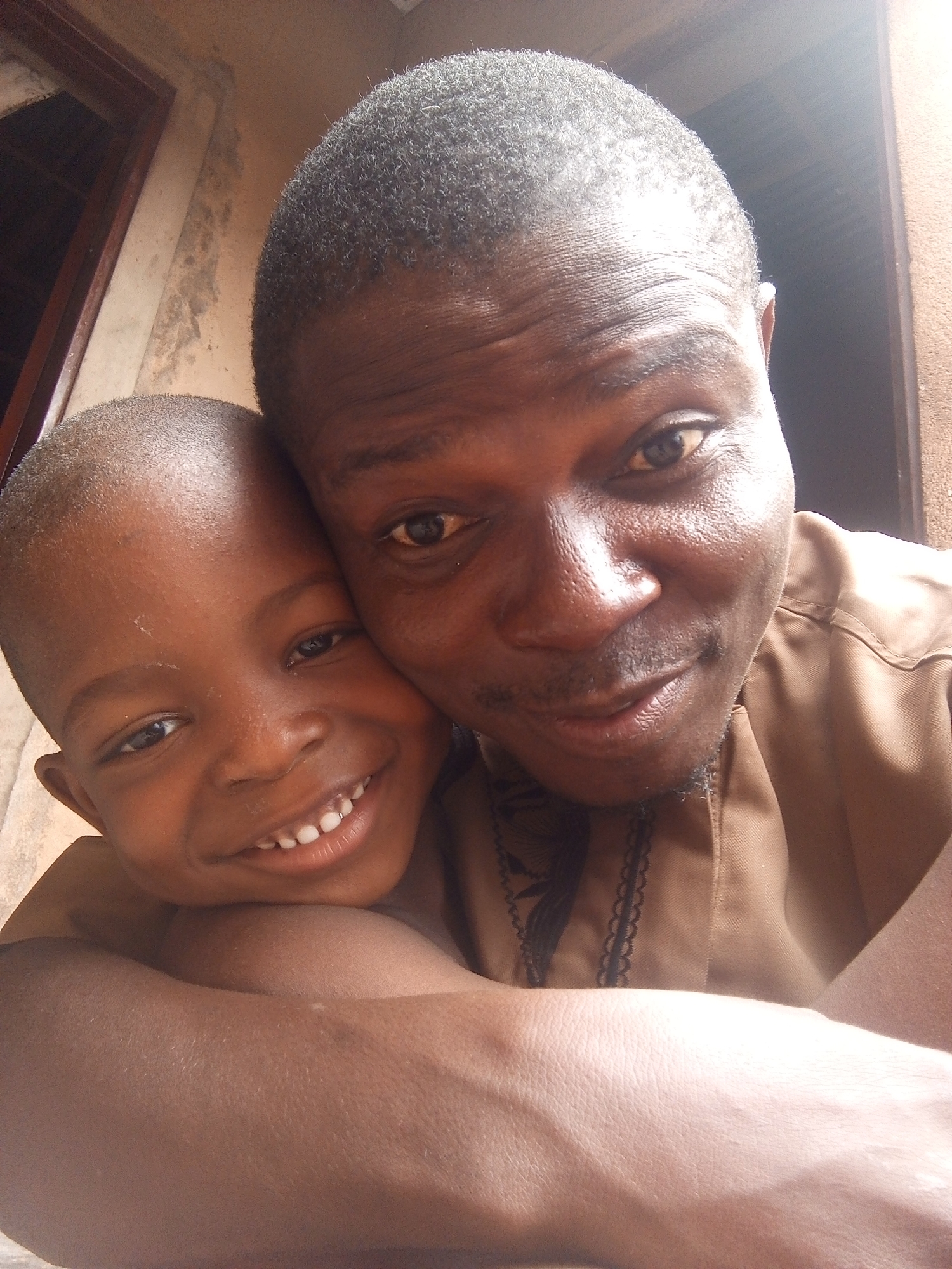 A close-up selfie captures a dark-skinned man and a young boy embracing. The man is wearing a brown shirt with decorative trim around the collar. The background shows part of a beige wall with a dark wooden framed window. The location appears to be Tempure, Nigeria.