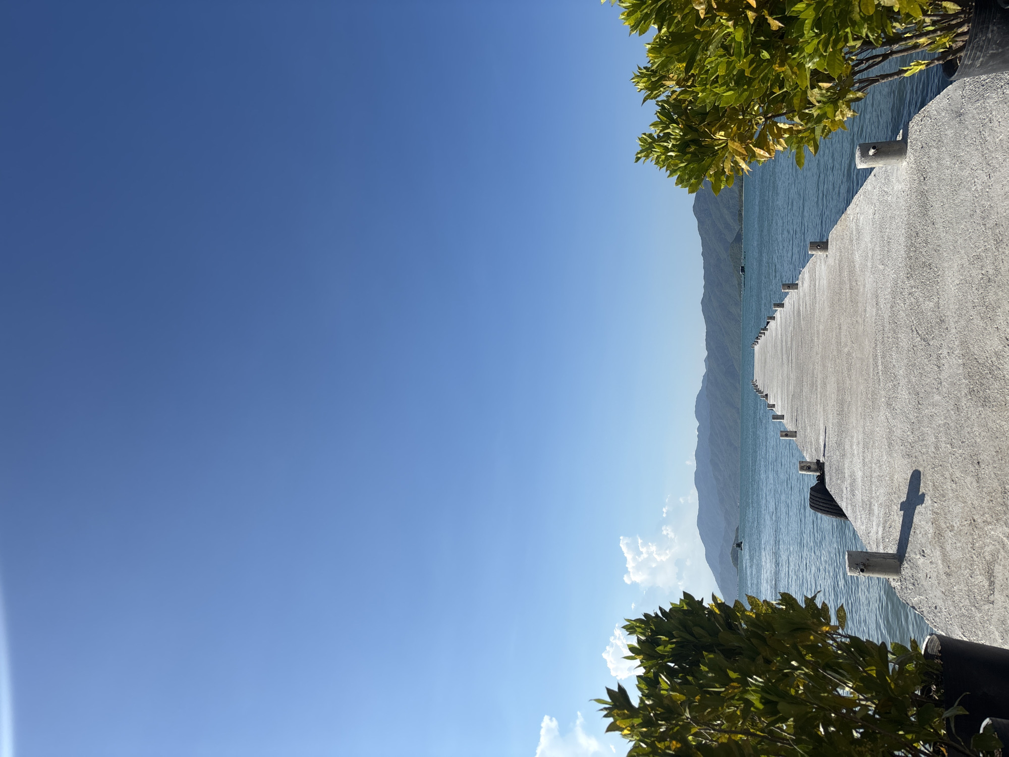 An extended concrete pier stretches into a calm, blue body of water under a clear, bright blue sky. The pier is lined with small, cylindrical concrete bollards and a single spare tire is visible near the start of the pier. Lush green foliage in large black pots frames the left and right sides of the pier's foreground. In the distance, rolling green mountains form a backdrop to the water. A small boat is visible on the water between the pier and the distant mountains, and a few distant structures, possibly fish farms, are also present in the water. The scene depicts a serene waterfront location in the Vĩnh Lương commune of Nha Trang city, Vietnam, likely a public access point or a part of a resort.