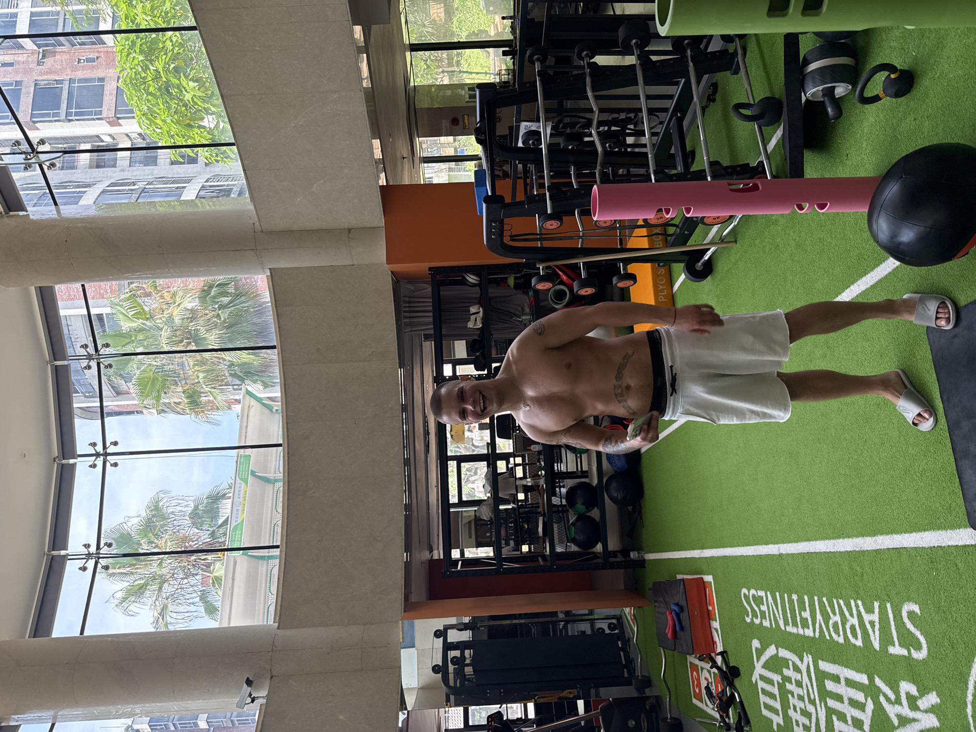 The image depicts a male individual posing inside a modern gym identified as "Starry Fitness" in Haizhu District, China.
The central figure is a muscular man with a lean physique, standing shirtless and facing the camera with a wide smile. He has short, light-colored hair, and tattoos are visible on his right shoulder/bicep and across his abdomen, below the navel. He wears white athletic shorts and grey slide sandals. In his left hand, he holds a green smartphone. A thin silver chain with a pendant is around his neck.
The gym features a green artificial turf floor with white markings and the white Chinese characters "星悦" (Xīng Yuè) and the English text "STARRYFITNESS" emblazoned on it. Various gym equipment is scattered across the floor, including black medicine balls, kettlebells, weight racks with orange-rimmed plates, an ab roller, a pink cylindrical stand, and a "PLYO S BOX." In the background, additional weight machines and racks are visible.
The gym is characterized by large, curved floor-to-ceiling windows supported by a thick, light-colored marble column. Through these windows, an urban exterior is visible, featuring multi-story buildings constructed from brick and concrete, interspersed with numerous palm trees and other green foliage. The sky outside is clear and blue. A white security camera is mounted on the large column within the gym, facing the workout area. A green sign with white Chinese characters is visible on a building through one of the windows.