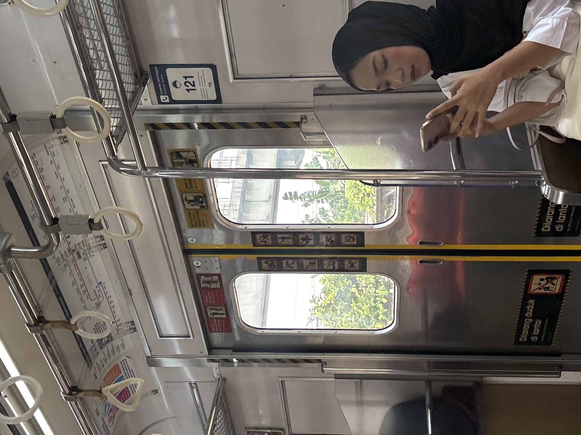In an indoor public transport setting, a young woman wearing a black hijab and a white shirt is seated, looking down at her rose-gold smartphone. The train car interior features metal doors with windows, handrails, and various signs. A blue sign with the number "121" is visible on the right, and signs with pictograms and text in Indonesian, such as "Dilarang duduk di lantai" (Do not sit on the floor), are present on the doors. Outside the windows, lush green foliage and a blurred overpass structure are visible, suggesting an urban environment. The overall lighting indicates it is daytime. The scene is consistent with travel on the MRT Jakarta system in North Jakarta, Indonesia.