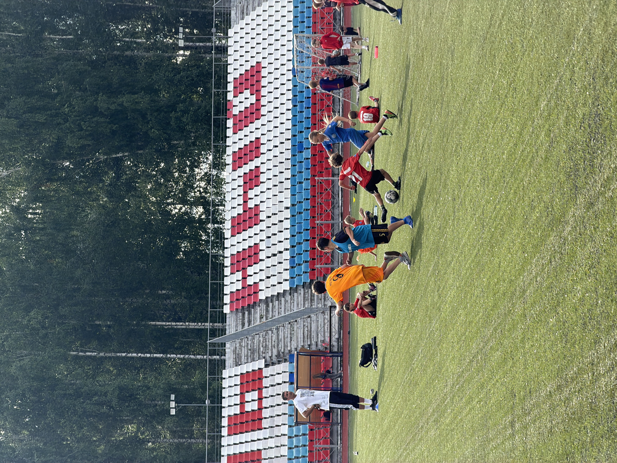 The image depicts a group of children and adults on a bright, sunny day at a soccer field in Chaikovsky, Russia. The scene is set against a backdrop of stadium seating with rows of red, white, and blue seats, and a dense forest of trees. The artificial turf of the soccer field shows clear markings for the game.
Several individuals, primarily young people dressed in athletic wear, are engaged in soccer-related activities. Some are actively playing with a soccer ball, while others appear to be practicing drills or warming up. One person in a blue jersey is kicking a ball, and another in an orange jersey is in motion. A young person in a red jersey with the number "19" is visible. Near the goalpost, other individuals are standing or moving.
In the foreground, a man in a white t-shirt and black shorts stands on the field, looking towards the left. A black soccer ball is on the ground near him. Further back, a black sports bag rests on the grass. The overall atmosphere suggests a training session or a casual game. The bright sunlight indicates it is likely daytime, and the clear skies suggest fair weather. The presence of a soccer field and stadium seating firmly places the scene in a sports venue. Some of the text visible on the jerseys is difficult to read clearly due to the image resolution and motion blur, but one jersey appears to have "19" and another has "5".