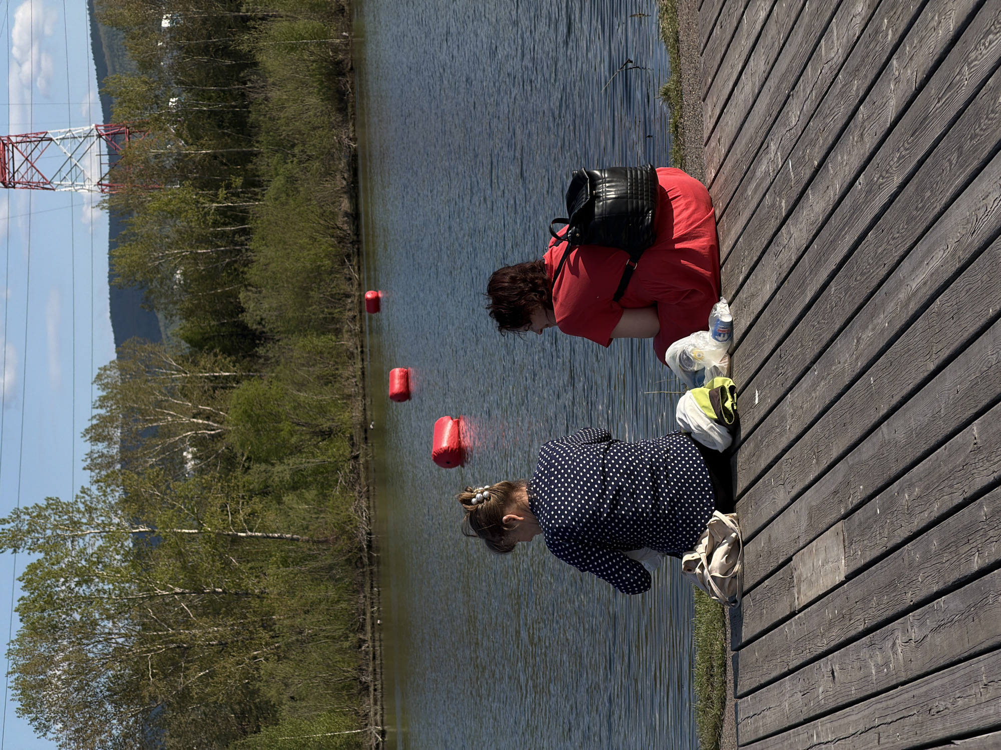Two individuals, an adult and a child, are crouched on a wooden boardwalk adjacent to a body of water, presumably a lake or river, marked with red buoys. The adult is wearing a red garment and a black backpack. The child wears a navy blue polka dot jacket. Debris, including a water bottle, plastic bags, and a green/yellow bag, are visible next to the adult. The location is in Krasnoyarsk, Russia, as indicated by a red and white power-line tower and the foliage along the waterfront, which are typical of that region.