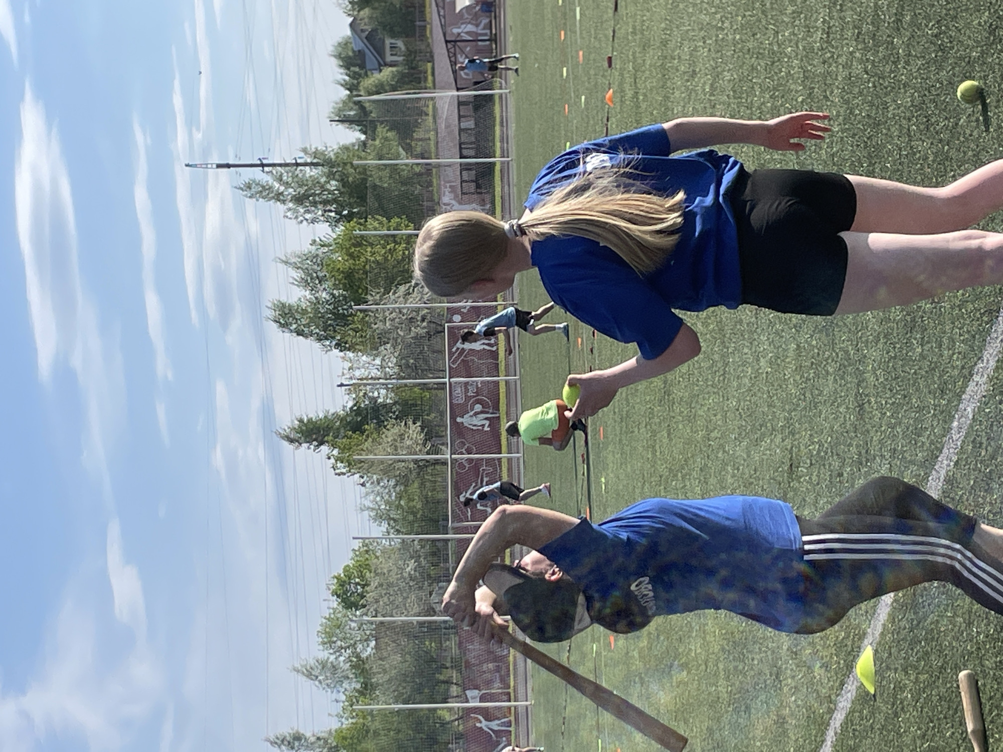 The image shows two individuals on a grass field under a sunny sky, possibly in Zav'yalovo, Russia, based on visual features. One individual, a girl, is wearing a blue shirt and black shorts and is holding a tennis ball. The other individual is wearing a blue shirt and dark pants with stripes and is holding a baseball bat. In the background, there are other people, cones, a chain-link fence, trees, and a building.