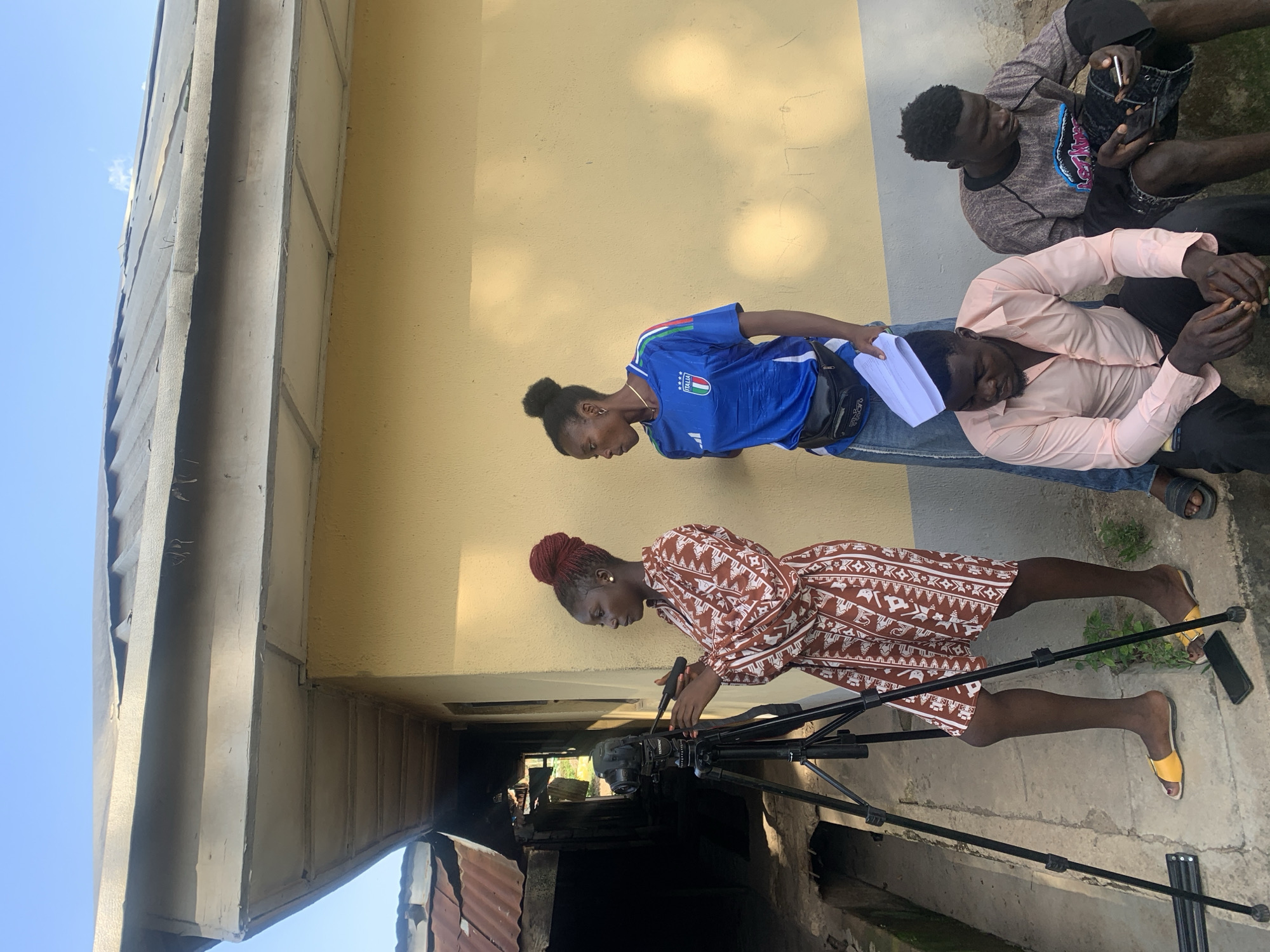 An outdoor scene in Jos, Nigeria, features four individuals engaged near a single-story building under a clear blue sky. A woman with red braided hair, wearing a brown and white patterned dress and yellow sandals, stands on the left, operating a black camera mounted on a tripod. A black smartphone rests on the concrete ground near her feet. To her right, a second woman, wearing a blue "ITALIA" branded t-shirt, denim skirt, and a black fanny pack, stands with white papers in her left hand, observing. In the foreground, two men are seated on a concrete ledge. The man on the left wears a light pink button-up shirt and dark trousers, looking downwards. Beside him, a second man wears a grey and black patterned long-sleeved shirt and dark shorts, appearing to interact with a small device in his hands. The building behind them has a light yellow stucco-like exterior wall and a grey metal roof with a white soffit. To the far left, an older, darker structure with rusty corrugated metal roofing is visible, along with a narrow pathway or drainage channel.
