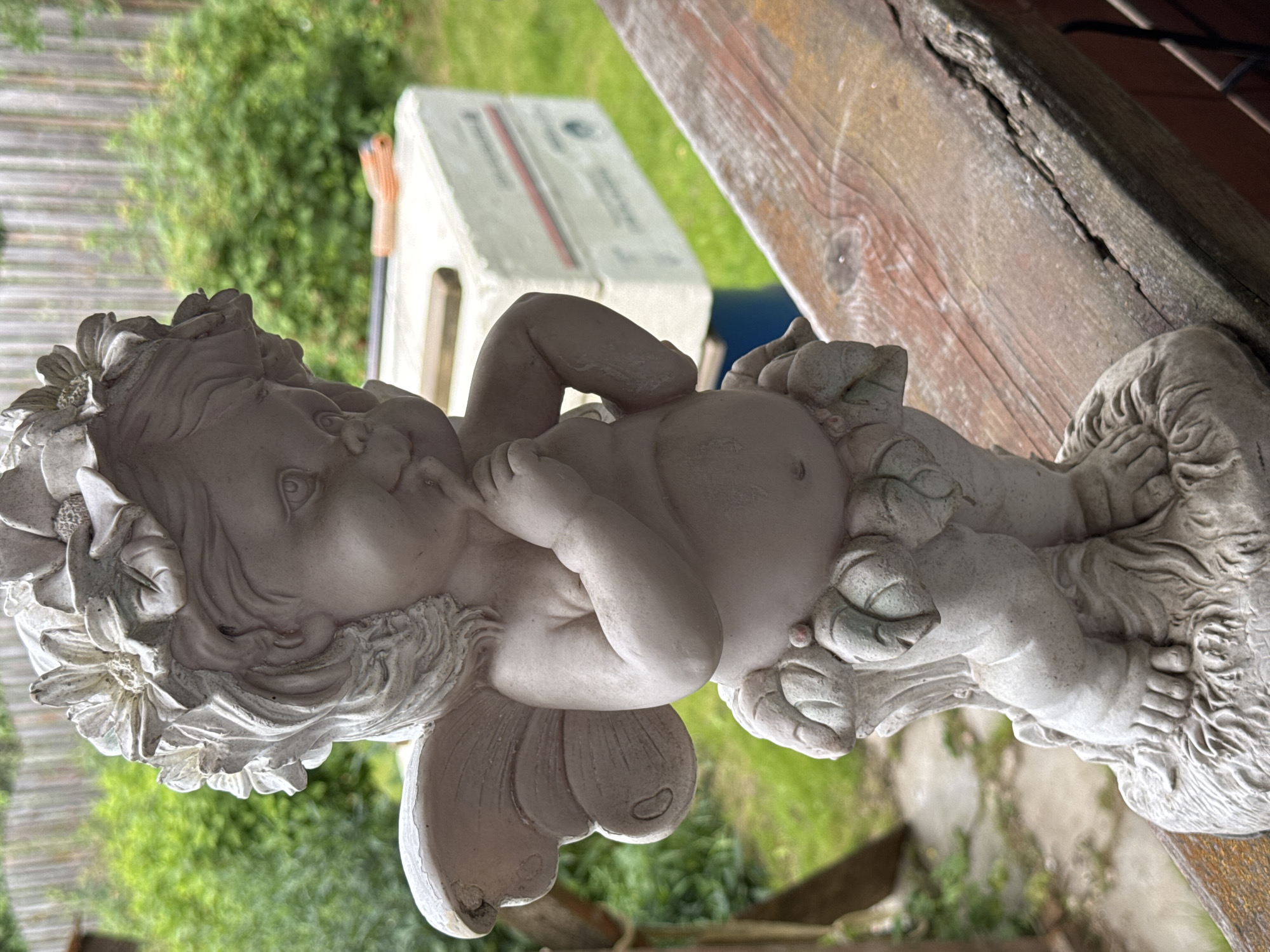 A light pink, weathered statue of a cherub or fairy stands on a weathered wooden railing. The statue is of a chubby child with delicate wings, adorned with a floral wreath on its head, and appears to be in a thoughtful pose with a finger to its chin. The background is blurred, showing greenery and a wooden fence, suggesting an outdoor setting, possibly a garden or backyard. In the middle ground, a white object, possibly a cooler or storage container, is visible with some tools resting on it. The scene appears to be located in Bersenevka, Russia.