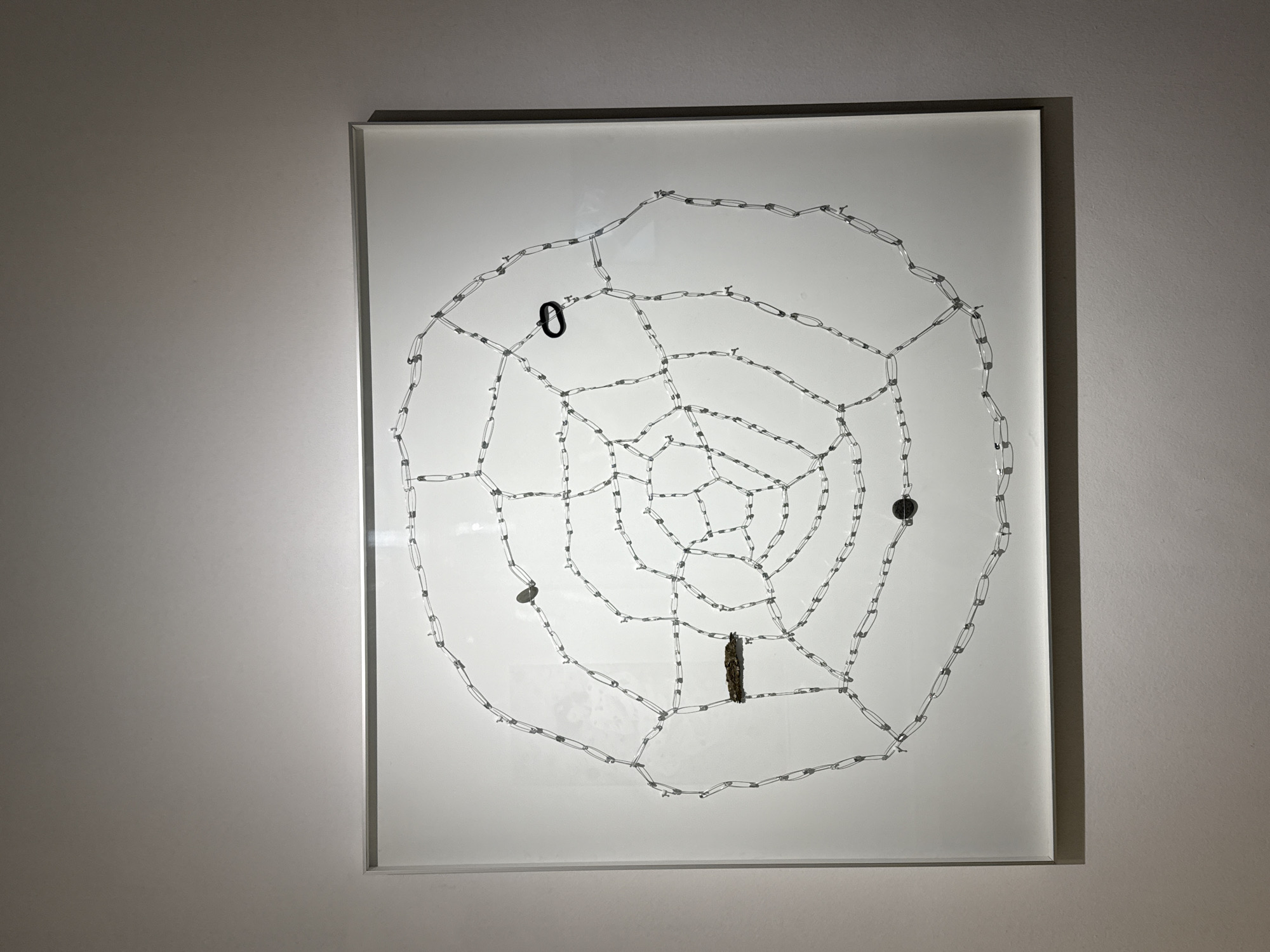 A framed artwork hangs on a light gray wall. The artwork depicts a stylized spiderweb constructed from a chain-like material connected by small, cylindrical elements, with a white background. The spiderweb is not perfectly symmetrical. Four small, dark objects of varying shapes are affixed to the web: a small, black, looped object, a small, round, flat object, and a cylindrical, textured object. The artwork is encased in a light gray frame. The photograph appears to have been taken indoors. The location is not discernible from the image.
