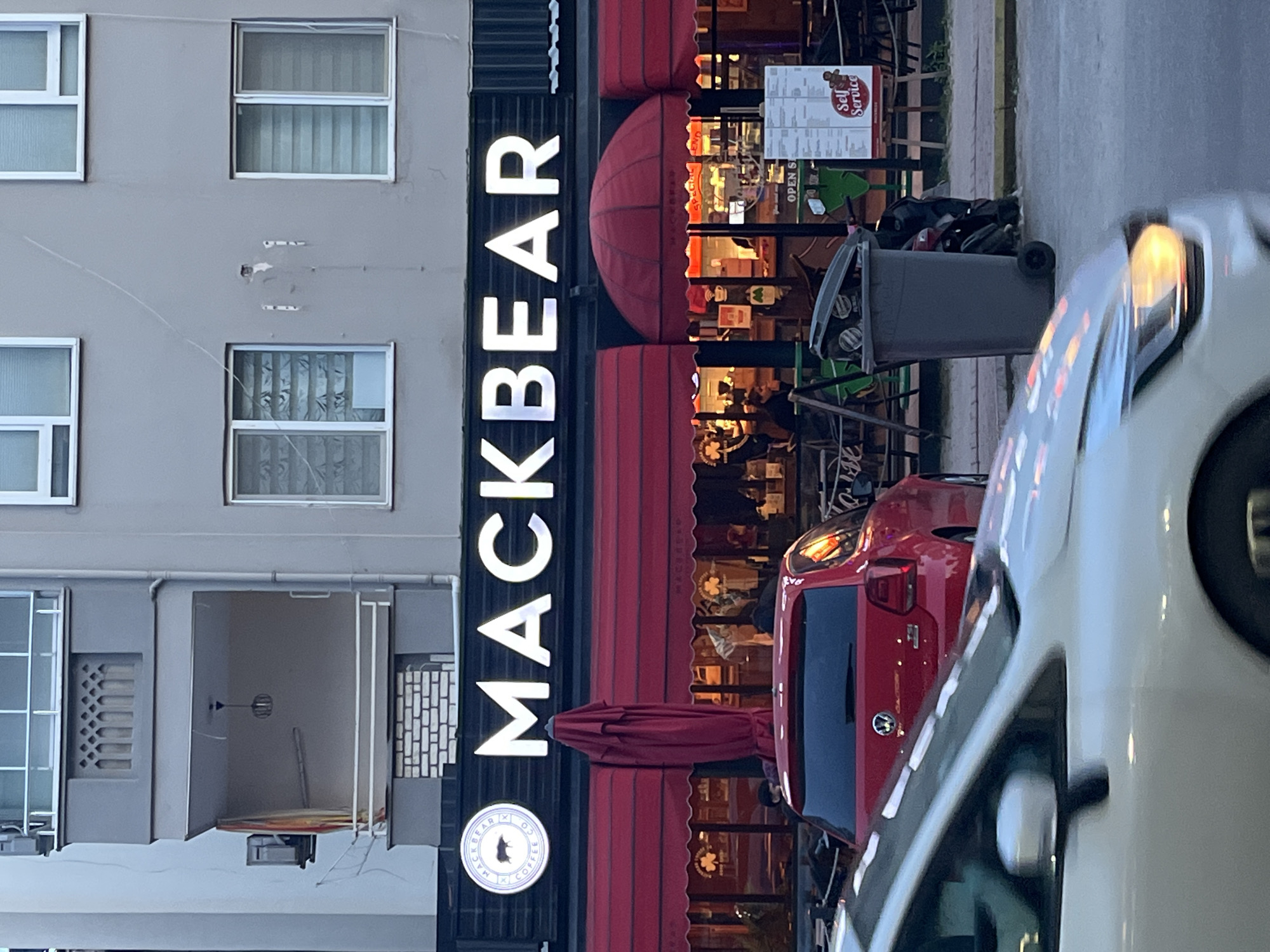 The image depicts the front of a café or restaurant named "MACKBEAR" located in Alanya, Turkey. The building has a gray facade with multiple windows. A vertical sign with the café's name is prominently displayed on the right side of the building. Red awnings cover the entrance and outdoor seating area. Inside, people can be seen seated and enjoying the café's atmosphere. A menu board offering "Self Service" options is placed near the entrance. A trash can and parked cars partially obscure the view. The scene suggests an urban setting, possibly a street or commercial area within Alanya.