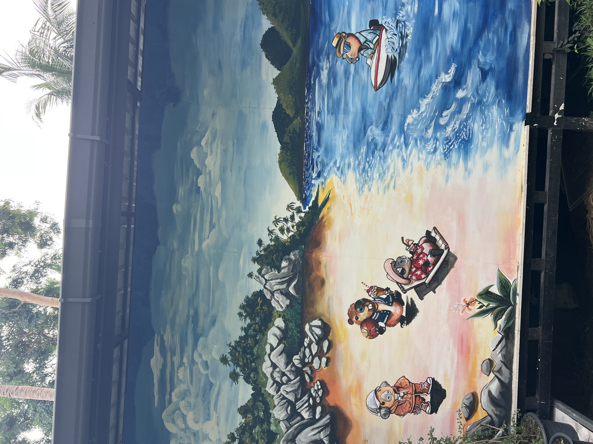 The image shows a large mural on a wall, likely outdoors, in Baan Lamai, Thailand. The mural depicts a beach scene with cartoon animals. In the top right, one animal is surfing. Below, on the beach, two animals are in a beach chair, another animal is walking with an ice cream and basketball, and another is dressed as an astronaut. The background includes a blue sky with clouds on the left and a mountain range in the distance on the right. Palm trees line the beach. Rocks and tropical plants are painted in the foreground. The mural is painted on what appears to be several joined boards, supported by a black metal frame. Part of a building or shelter structure is visible above and to the left of the mural, including a palm tree. Vegetation is visible to the right of the mural.