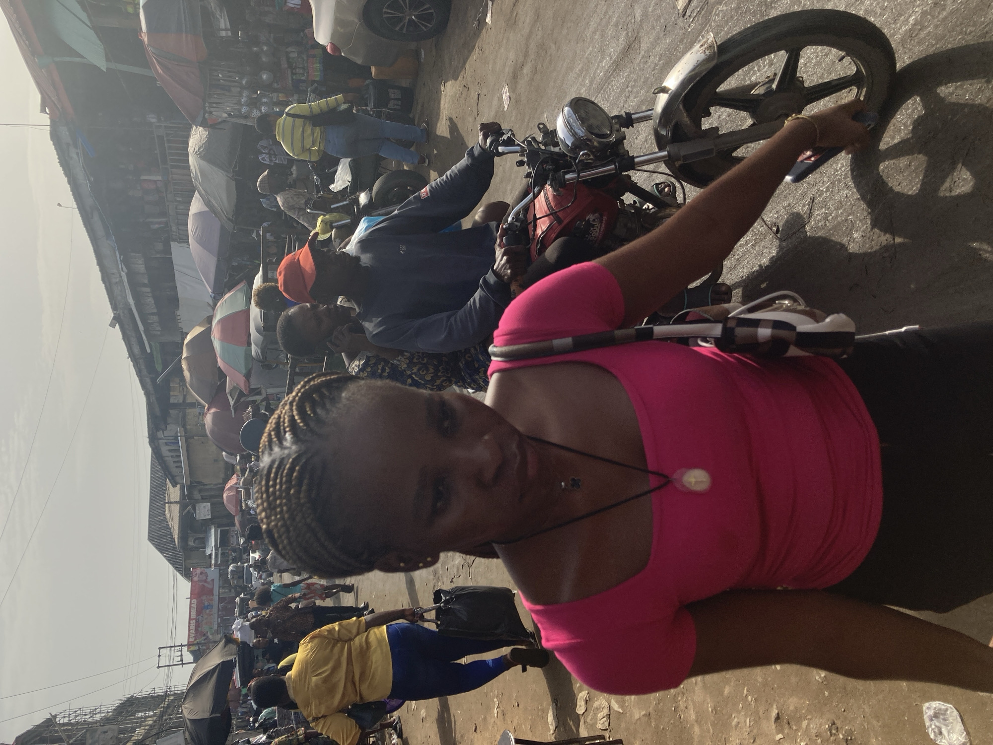 The image depicts a bustling street scene in Ura Nta Umuarandu, Nigeria. The foreground is dominated by a woman wearing a pink top and dark pants, with her hair braided. She appears to be near or on a motorcycle. The motorcycle, partially visible, is red with a silver front wheel. Behind her, a man in a dark jacket and orange cap is seated on the motorcycle as well, possibly the driver. Further in the background, a crowd of people can be seen, some holding umbrellas. A building and various signs are visible in the background. The street surface is rough and uneven. The overall impression is of a crowded and active urban environment.