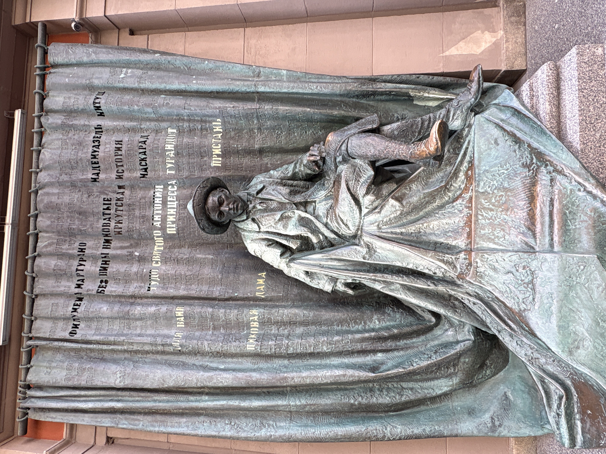 This is a full-shot, eye-level, medium-format photograph of a bronze sculpture in Moscow, Russia. The sculpture depicts a seated man in a hat and coat, draped in fabric that resembles a curtain. The man is positioned in front of a large, curtain-like bronze backdrop with engraved text. The text appears to be titles of literary works or plays, some of which are highlighted in gold lettering. The sculpture is situated outdoors, with a building facade visible in the background. The overall tone of the image is somber and artistic.