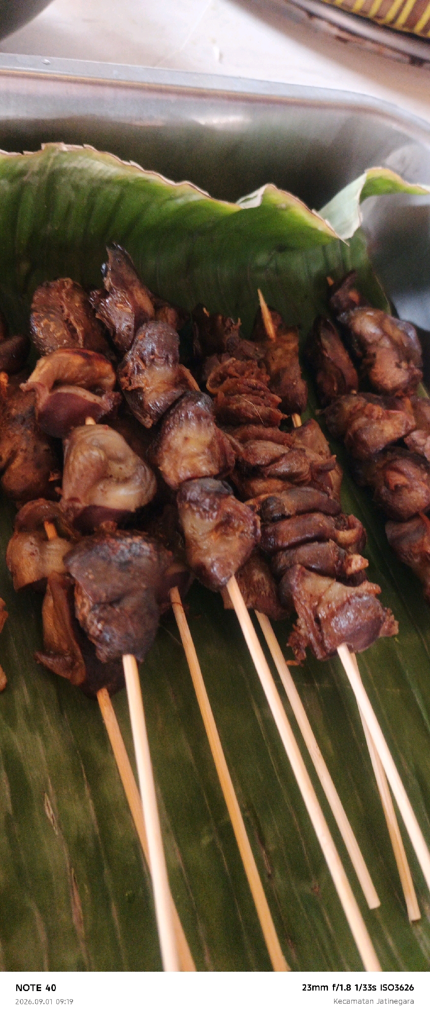 A close-up, high-angle shot displays several skewers of cooked chicken offal arranged on a bed of large, green banana leaves. The offal pieces are dark brown and appear to be grilled, with some showing a glistening texture. Wooden skewers pierce through the meat, with the ends of the skewers extending upwards. The leaves are large and have prominent veins. The bottom of the image contains text identifying the device as "NOTE 40", the date and time as "2026.09.01 09:19", the camera settings as "23mm f/1.8 1/33s ISO3626", and the location as "Kecamatan Jatinegara", which is a district in East Jakarta, Indonesia.