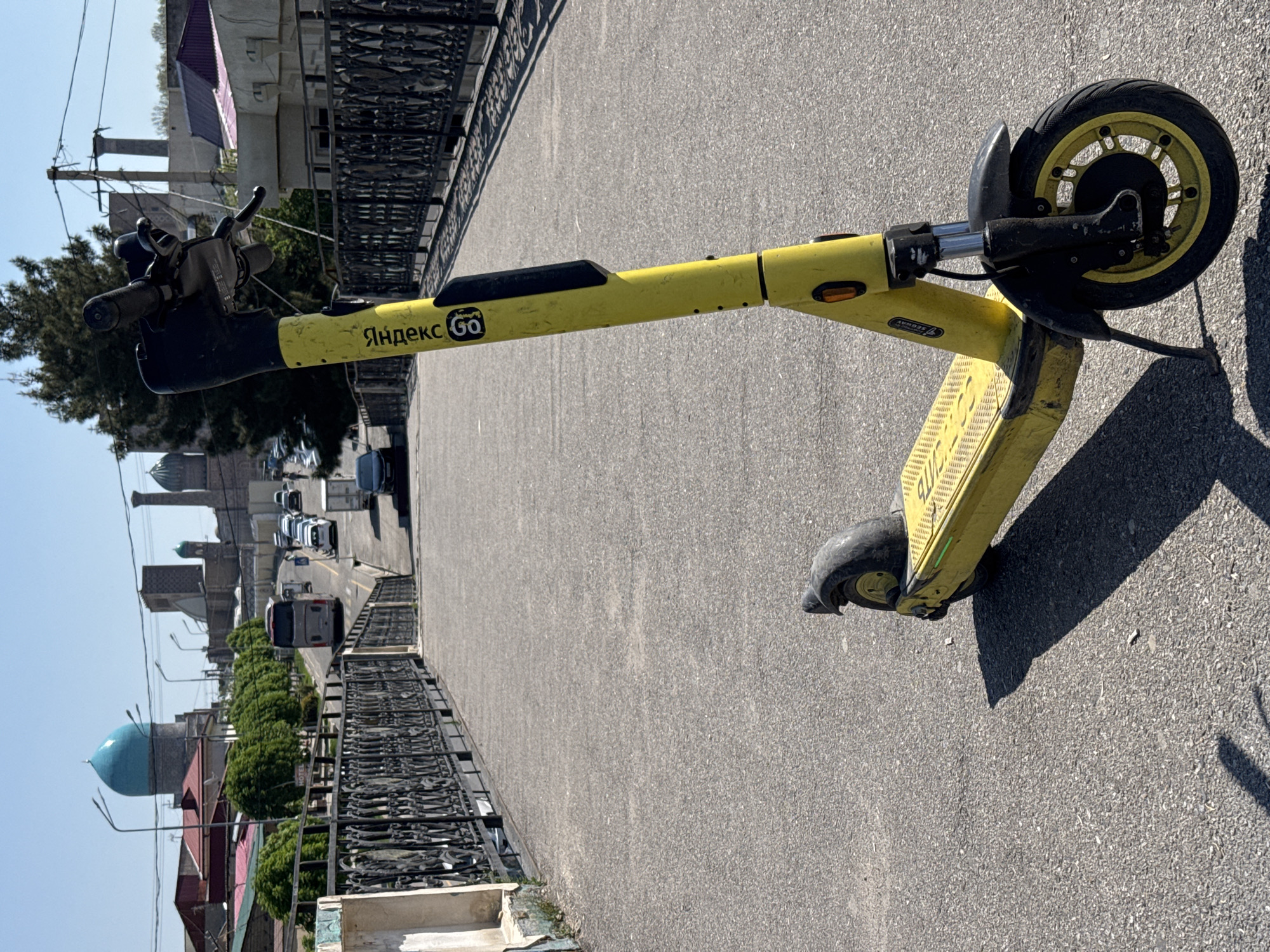 A yellow Yandex Go electric scooter is parked on a gray paved surface, likely a sidewalk or bridge, in Samarkand City, Uzbekistan. The scooter is oriented diagonally across the frame, with the front wheel to the right. A decorative black metal railing lines the left side of the paved surface. Beyond the railing is a street with cars and a bus. In the background are buildings, trees, and a mosque with a blue dome. The sky is blue. The lighting suggests a sunny day.