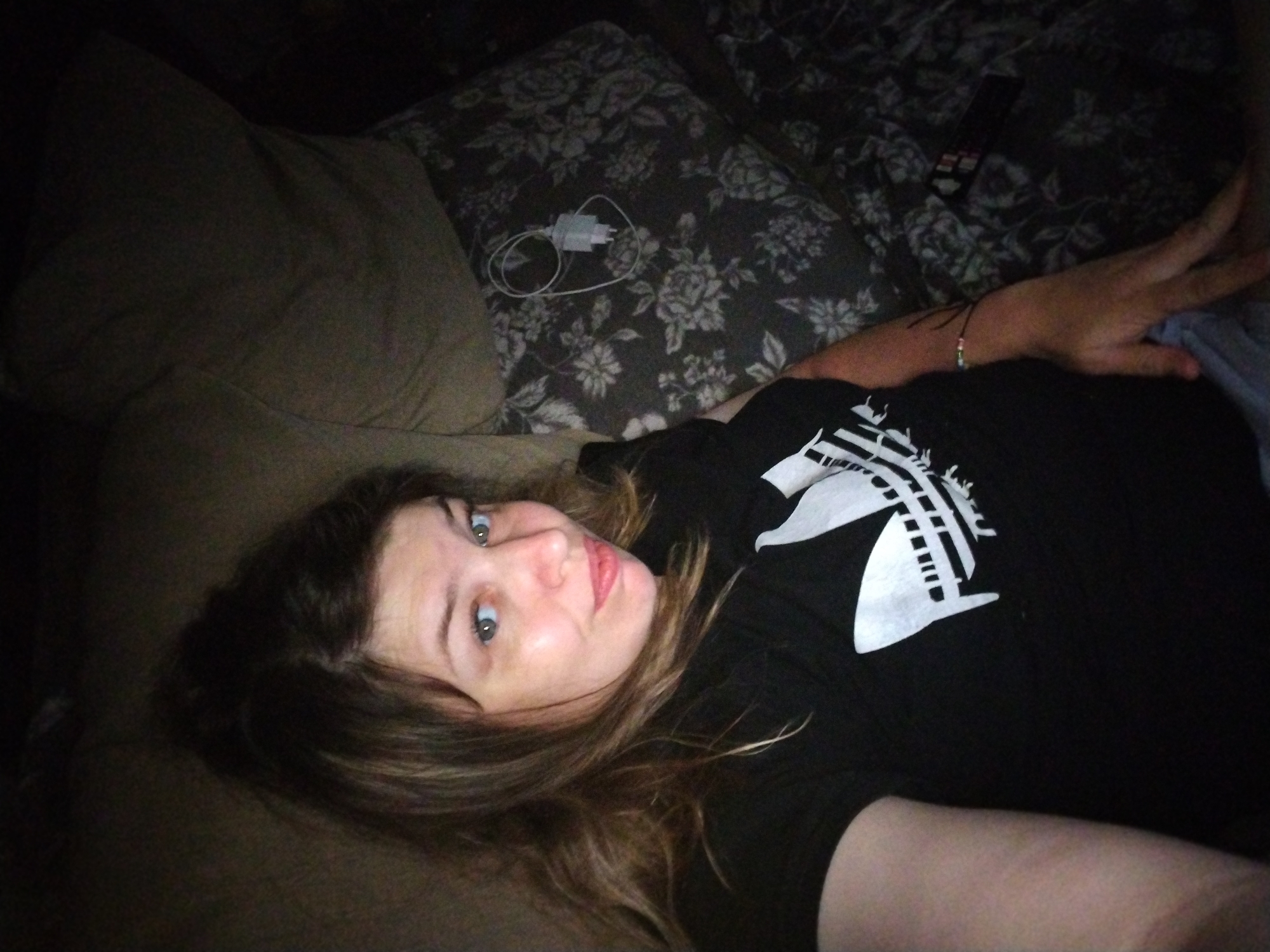 A young woman is lying on a couch, looking at the camera. She has long, brown hair and blue eyes. She is wearing a black t-shirt with a white graphic on it. A white phone charger is on a floral patterned cushion behind her. The room appears to be dimly lit.