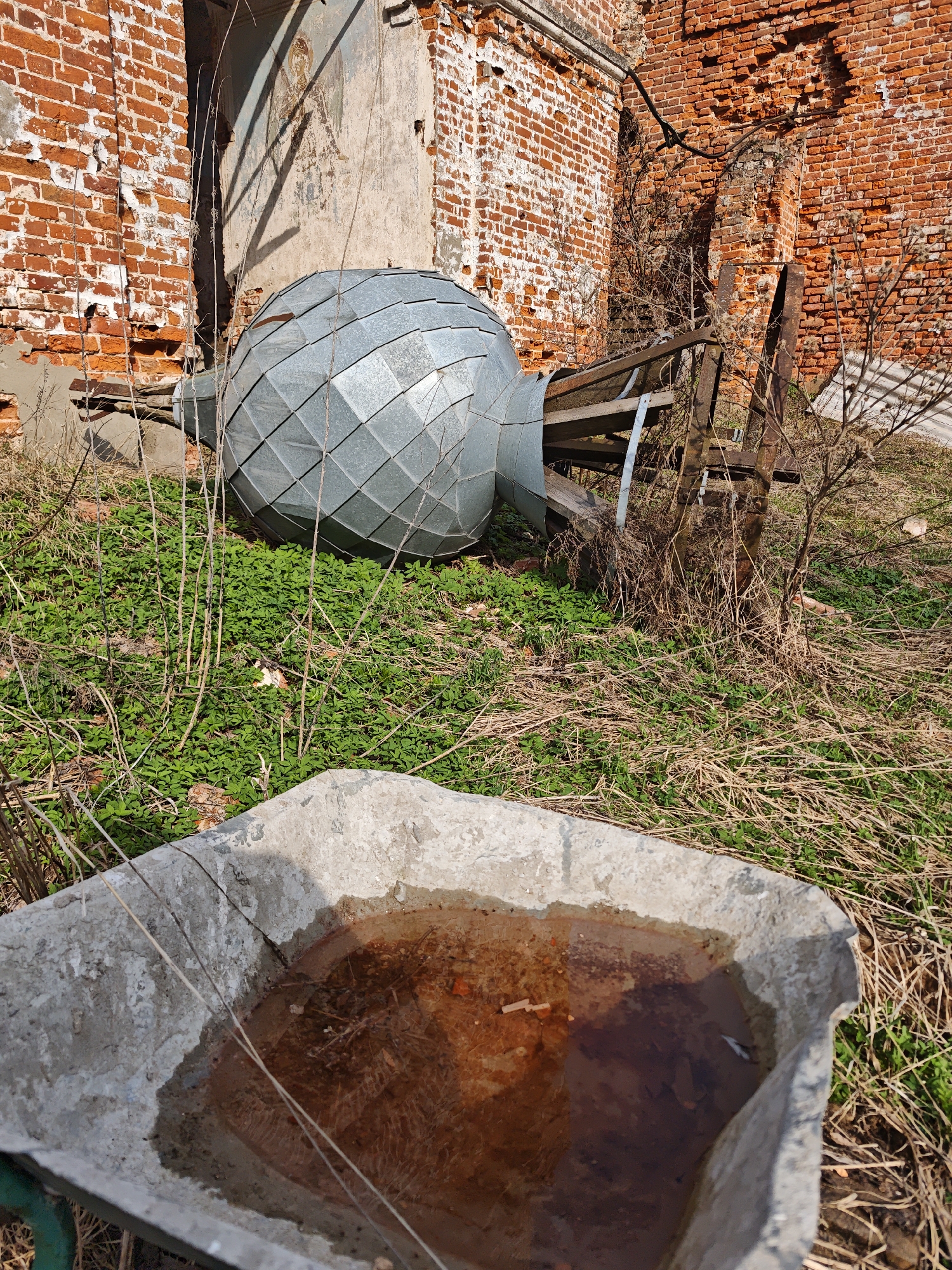 The scene depicts the grounds of what appears to be a dilapidated brick structure, possibly a former church, in the rural area of Ивановское (большерогачевский С/о), Russia. A large, metallic dome, likely part of a church cupola, lies on the ground amidst vegetation. Sections of the brick walls are crumbling, revealing the underlying structure. In the foreground, a concrete trough filled with brownish water sits amidst dry grass. A wooden structure lies near the metal dome. There is no visible human activity.