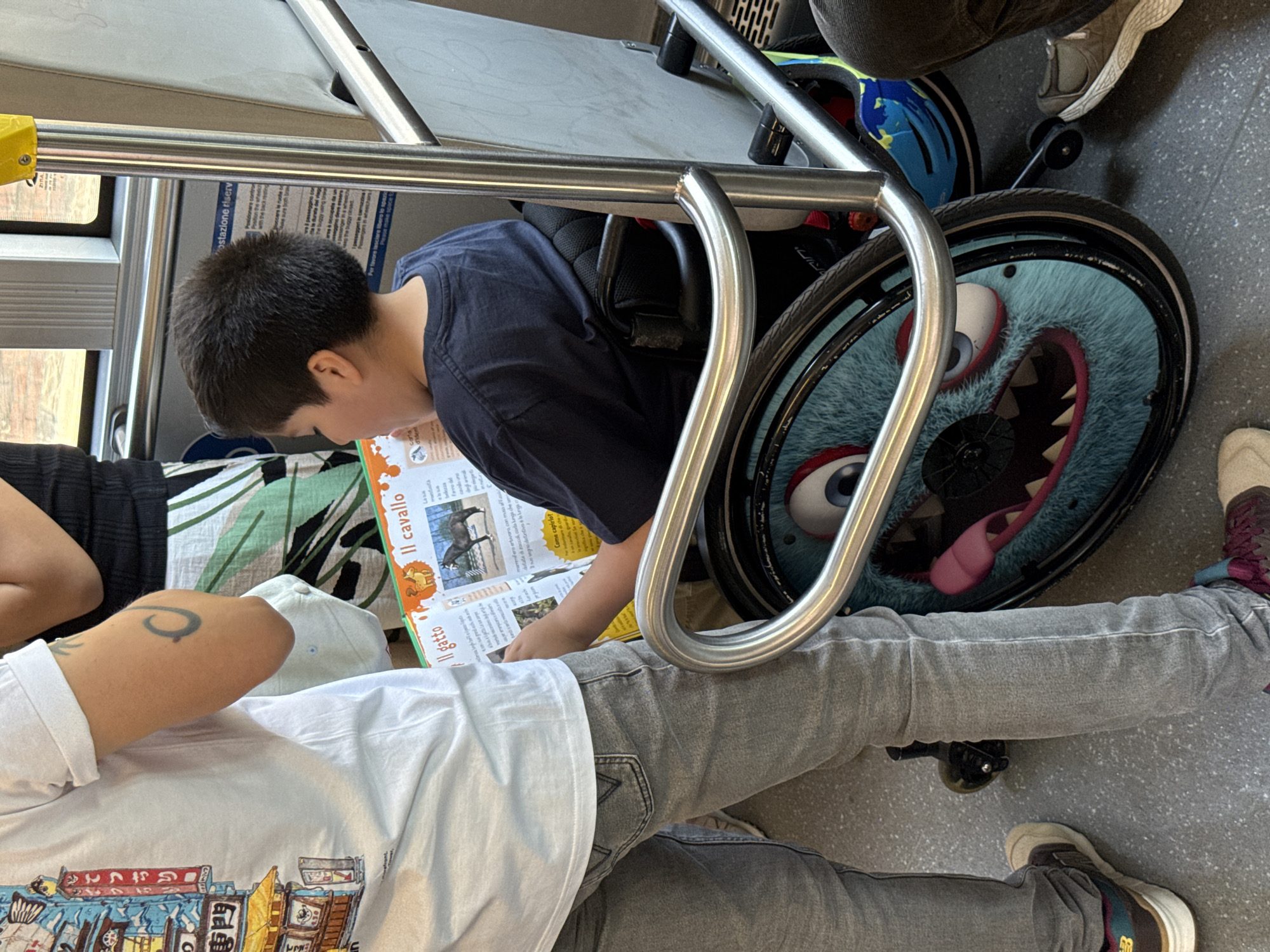 The image shows a person in a wheelchair on what appears to be a public transportation vehicle, potentially in Milan, Italy, given the Italian language text on the open book. The wheelchair wheel features a cartoonish monster design. A child is reading a book, and an adult is nearby. The surroundings suggest the interior of a bus or tram, based on the railings and seating arrangement.