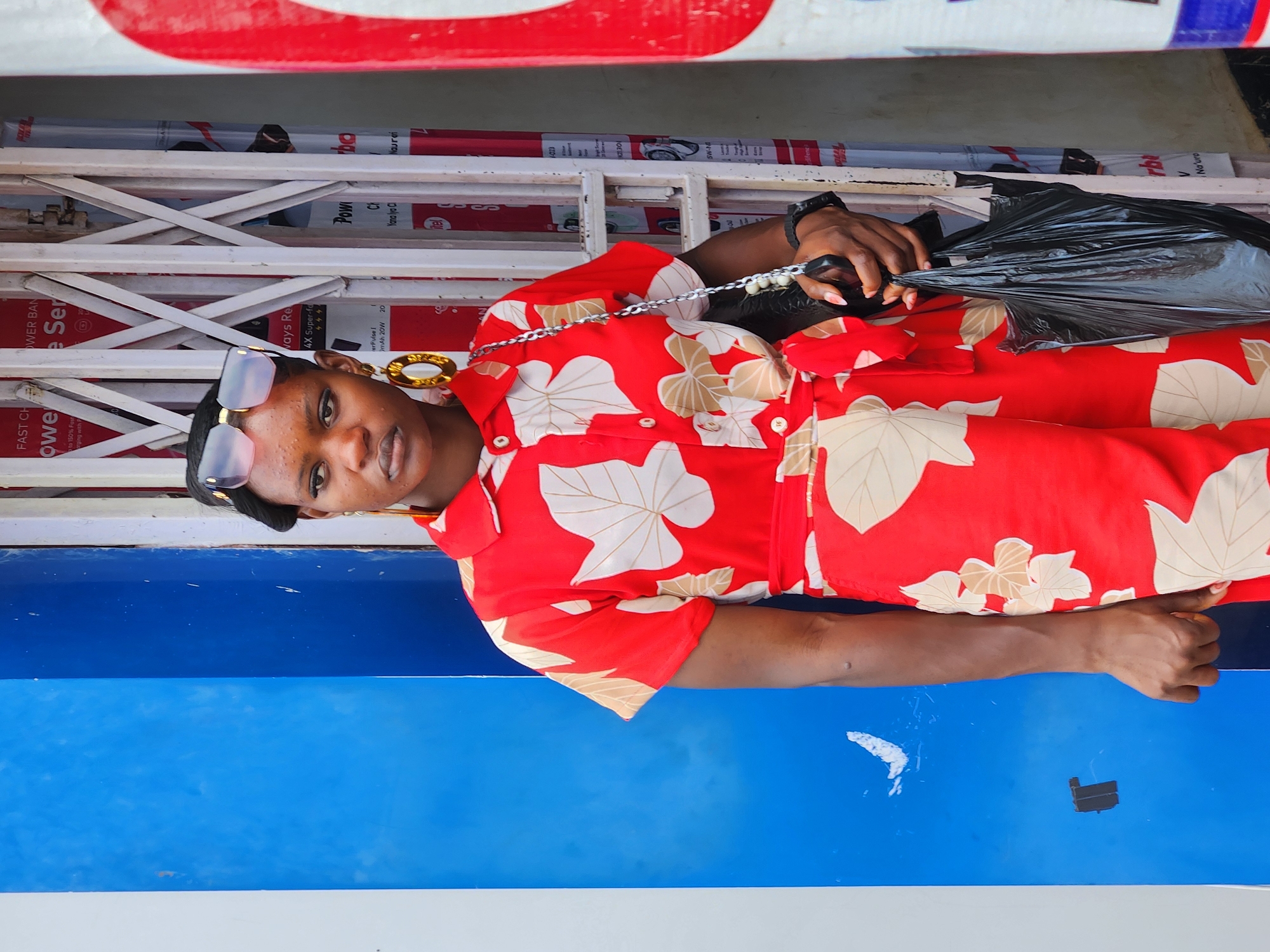 A person is posing for a photo, tilted on their side. They are wearing a red and white patterned shirt and sunglasses. They are holding a black plastic bag with their right hand, and a chain strap is visible over their left shoulder. A large gold hoop earring is visible on their left ear. The background consists of a blue surface and a metal gate with various signs visible behind it. Some of the visible text includes "FAST CH," "WER BAN," "eSer," "Ways Re," "4X Super-fo," "Pow," "CH," "er Pulse I," "mAh 20W 20," "Yana lya Cl," "Nouren," "1073," "21.30," "SW-18," and "rb." The lighting suggests it is daytime.