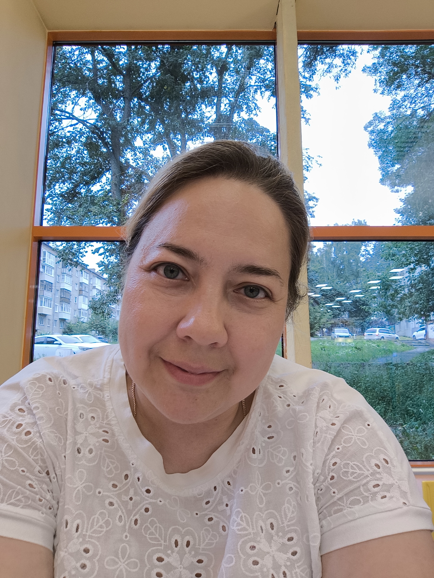 A woman is taking a selfie indoors, facing the camera. She is wearing a white eyelet shirt. Behind her is a window with a view of trees, buildings, and cars. The scene appears to be in Izhevsk, Russia, as indicated by the architecture and vegetation visible through the window. The light suggests it is daytime.