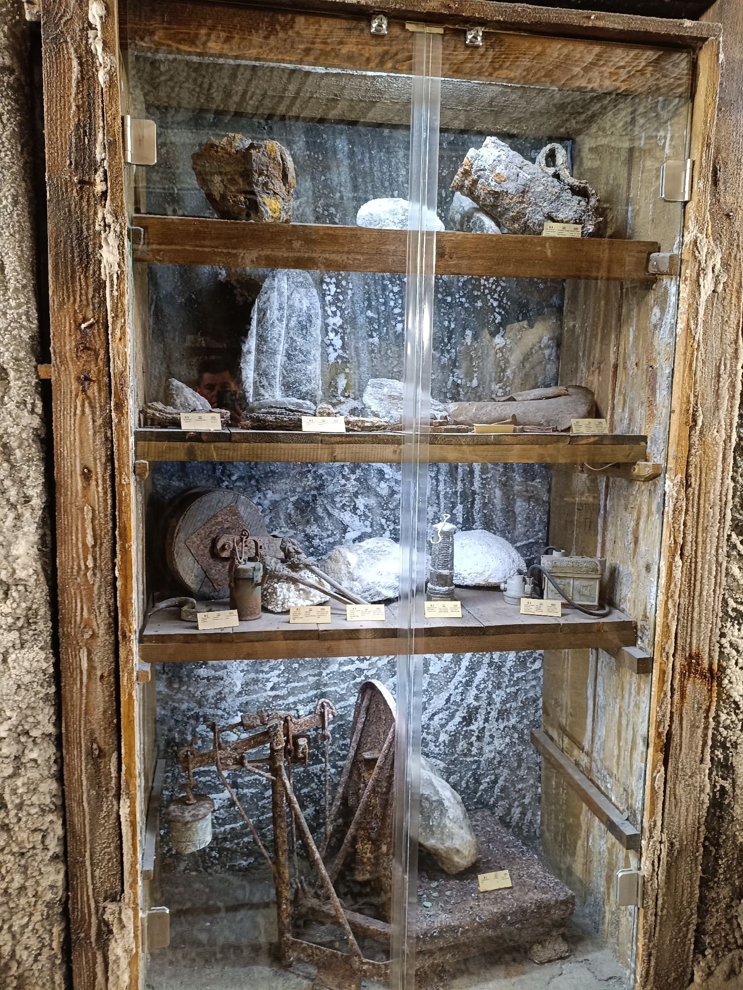 This media file shows a glass-fronted wooden cabinet displaying historical mining artifacts within a salt mine. The cabinet is filled with four shelves, each showcasing different items. The background walls appear to be made of rough, salt-encrusted rock, characteristic of a mine.
On the top shelf, there are several rock samples, some with yellow or brownish hues, and one with an attached metal loop.
The second shelf displays more rock samples and what appear to be primitive tools or geological specimens. There are also small informational placards with text.
The third shelf holds a large, rusty metal wheel, possibly part of a pulley system, along with other metal objects and rock samples. Again, informational placards are present.
The bottom shelf features a rusty weighing scale with a weight attached, and a large rock. Another informational placard is visible.
The overall impression is of a museum exhibit within an underground environment, likely a salt mine, preserved to showcase its history and the materials extracted. The lighting is somewhat dim, suggesting an indoor, subterranean setting. The salt formations on the wooden frame and the background walls are a prominent detail, indicating the unique location. No specific time of day or weather conditions are discernible. There are no people visible in the main frame, however, a reflection of a person taking the photo is visible in the glass on the second shelf. There are several small, yellow rectangular placards with text on them placed in front of most of the items.