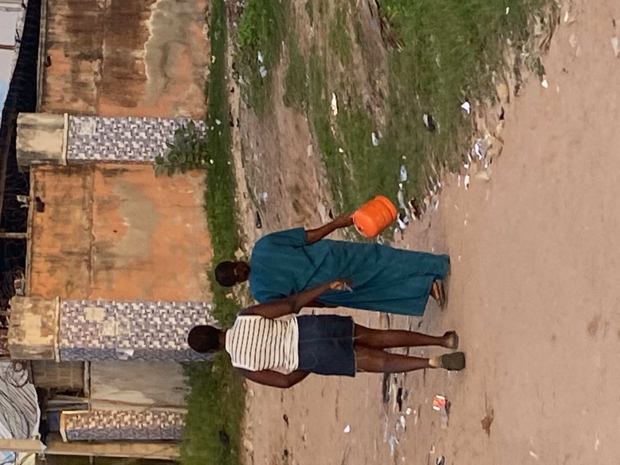 Two women are walking away from the camera down a dirt path littered with trash. The woman on the left wears a white and black striped sleeveless top and a dark blue denim skirt. The woman on the right wears a long, teal-colored dress and carries an orange bucket in her right hand. Behind them, a textured, orange-colored wall is visible, with decorative tiled columns on either side. The environment appears to be a dusty, somewhat neglected area.