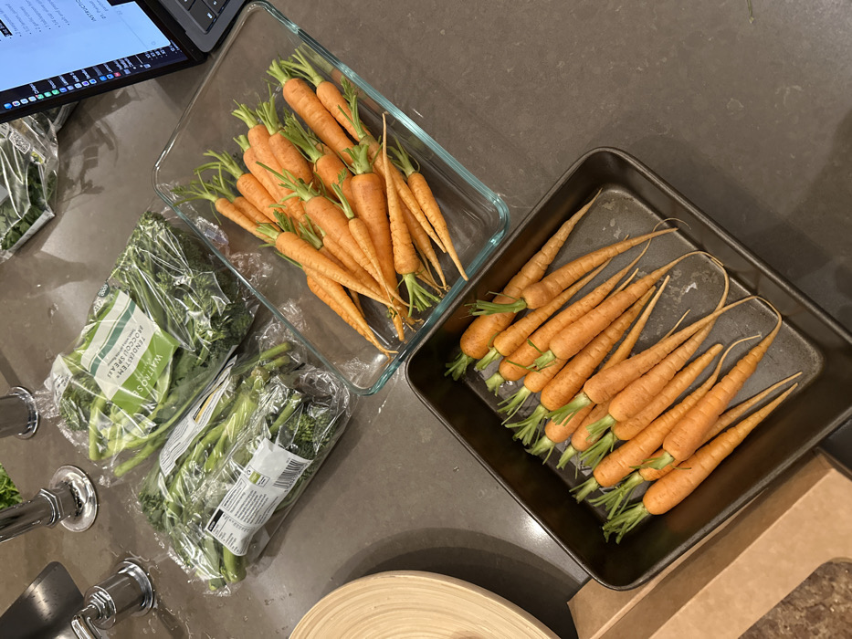 Two packages of tenderstem broccoli sit near a sink. Next to them are two containers filled with orange baby carrots with green tops. One container is clear glass, the other is dark metal. The ingredients sit on a dark gray countertop near a partially visible laptop with a recipe visible on the screen. A wooden bowl and cardboard box are partially visible on the lower right.
