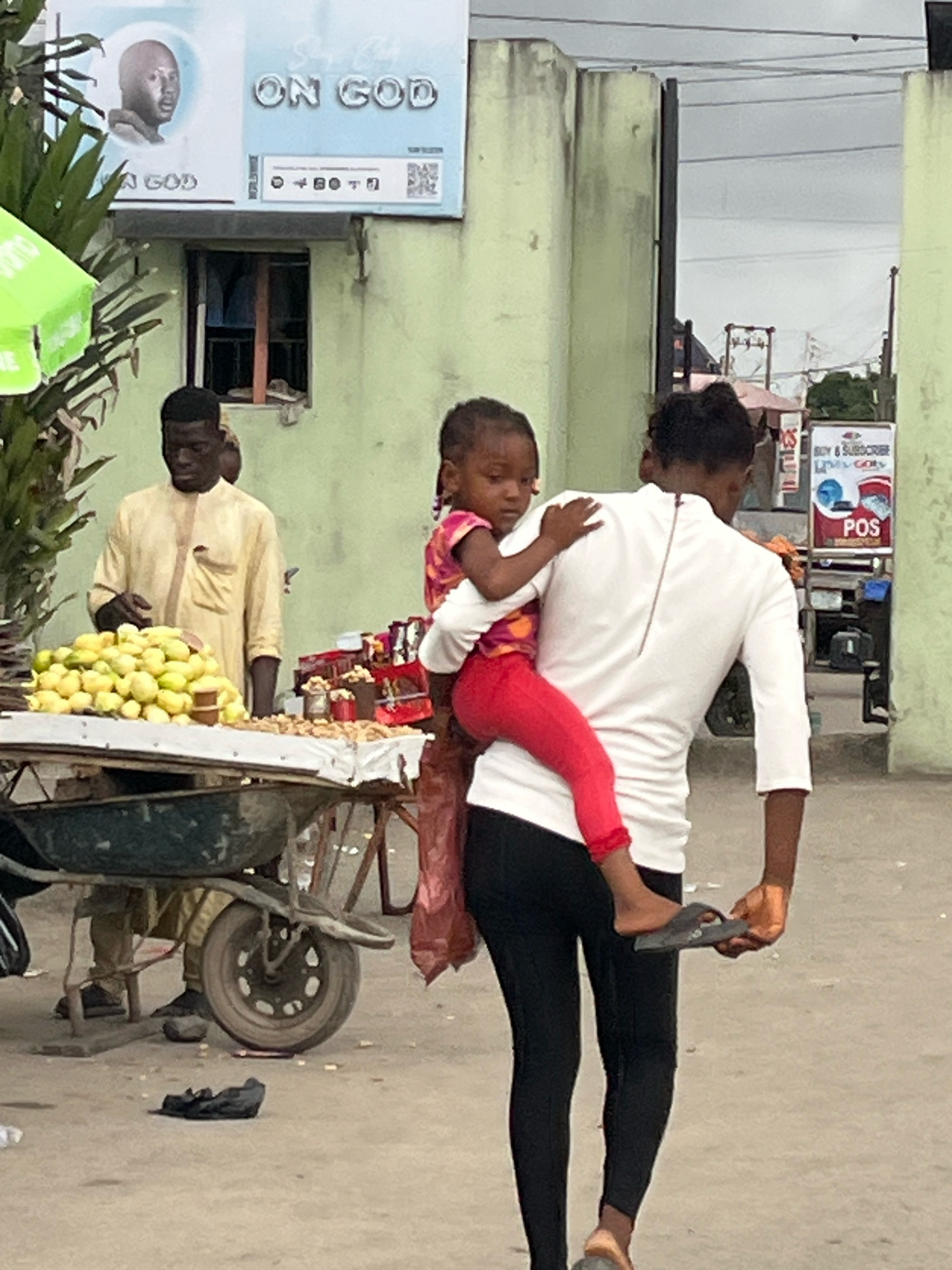 An outdoor scene in Ado, Nigeria shows a woman carrying a young child on her back. She is wearing a white shirt and black leggings. The child is wearing a pink and orange top and red leggings. The woman is holding a black flip-flop in her right hand. In the background, a man in a light yellow robe stands behind a cart laden with yellow fruits. A sign overhead reads "ON GOD." The environment appears to be a market or street scene.