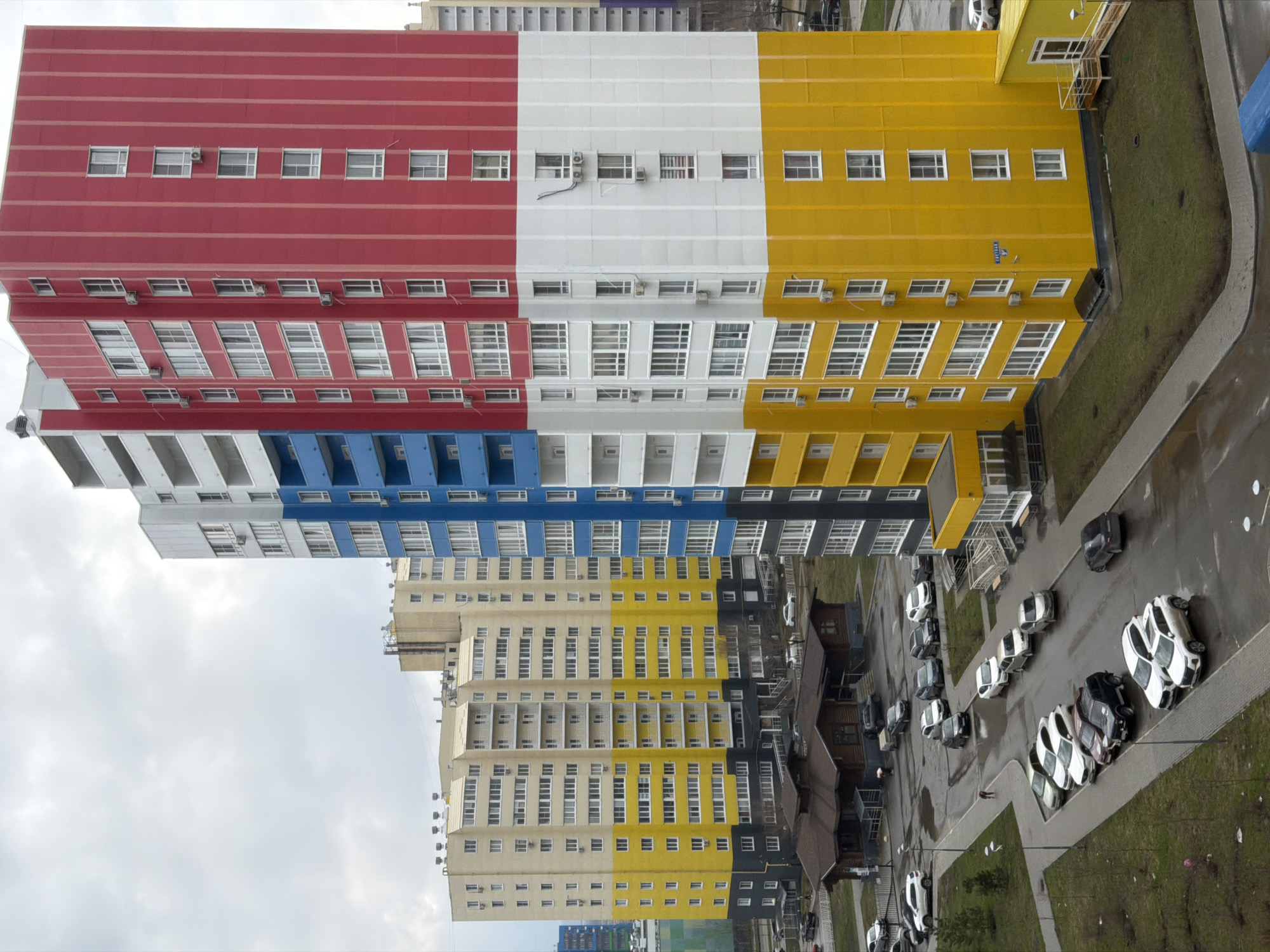 The image shows a view of a residential area in Засечное, Russia. Several high-rise apartment buildings dominate the scene, painted in a colorful pattern of red, white, blue, yellow, and beige. The buildings have rows of windows and balconies. A road runs alongside the buildings, with parked cars visible. There is also some green space with grass. The sky is overcast.