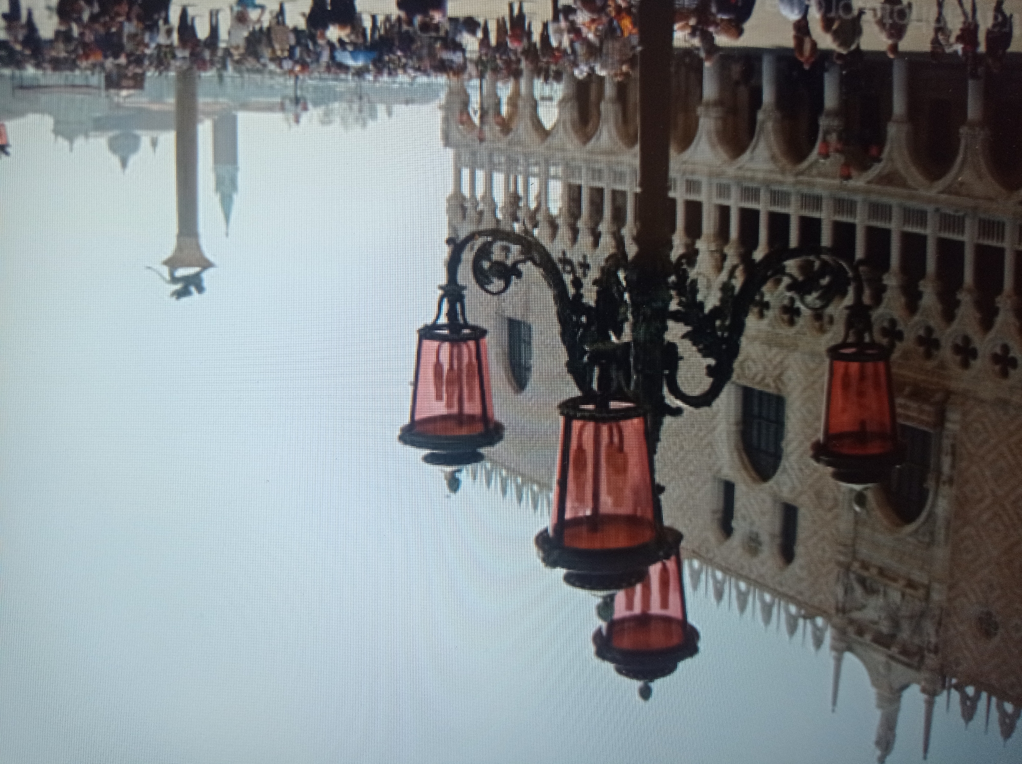 The image shows a wide shot of a large, ornate building with arched windows and decorative facades, possibly a palace or a government building. In the foreground, a multi-headed lamppost with red glass shades stands prominently. A crowd of people is visible in the square in front of the building. In the background, a tall column topped with a winged lion statue is visible, as well as a bell tower and domed buildings, suggesting a European city with historical architecture. The sky is bright and hazy. The image is grainy, suggesting it was taken with a lower-resolution camera or has been compressed.
This scene is not located in Mukachevo, Ukraine. The architecture strongly resembles Piazza San Marco in Venice, Italy, with the Doge's Palace on the left and St. Mark's Campanile and the Column of Saint Theodore in the background.