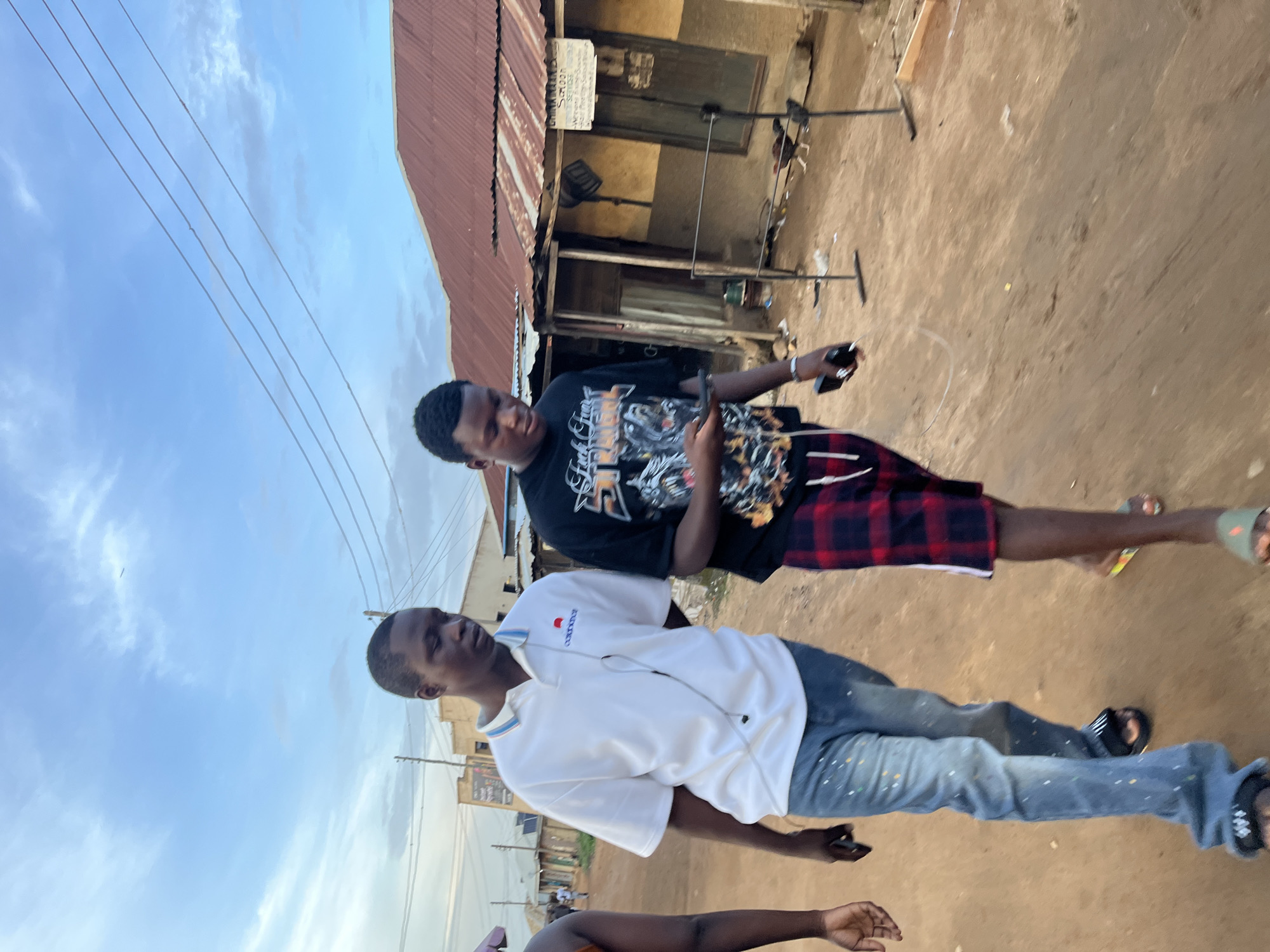 This image, set in Jos, Nigeria, captures two young men outdoors during what appears to be a bright day under a partly cloudy sky. The ground beneath them is unpaved, consisting of dirt or sand.
In the foreground, a man wears a white short-sleeved polo shirt, light blue jeans adorned with small colorful paint splatters, and black sandals. Earphones are visible around his neck, and he holds a smartphone in his left hand, looking downwards or away from the camera.
Behind him, another man is dressed in a black graphic t-shirt featuring text that appears to be "FUCK CRAP" alongside other designs, red and black plaid shorts, and light-colored sandals. He is actively engaged with a smartphone, holding it with both hands and focusing his attention on the screen. A third person's arm is partially visible at the bottom left of the frame.
The background features a simple building with a reddish-brown corrugated metal roof and worn, light-colored walls, suggestive of a local shop or residence. A blurry white sign is attached to the building. Multiple utility wires stretch across the sky above the scene.