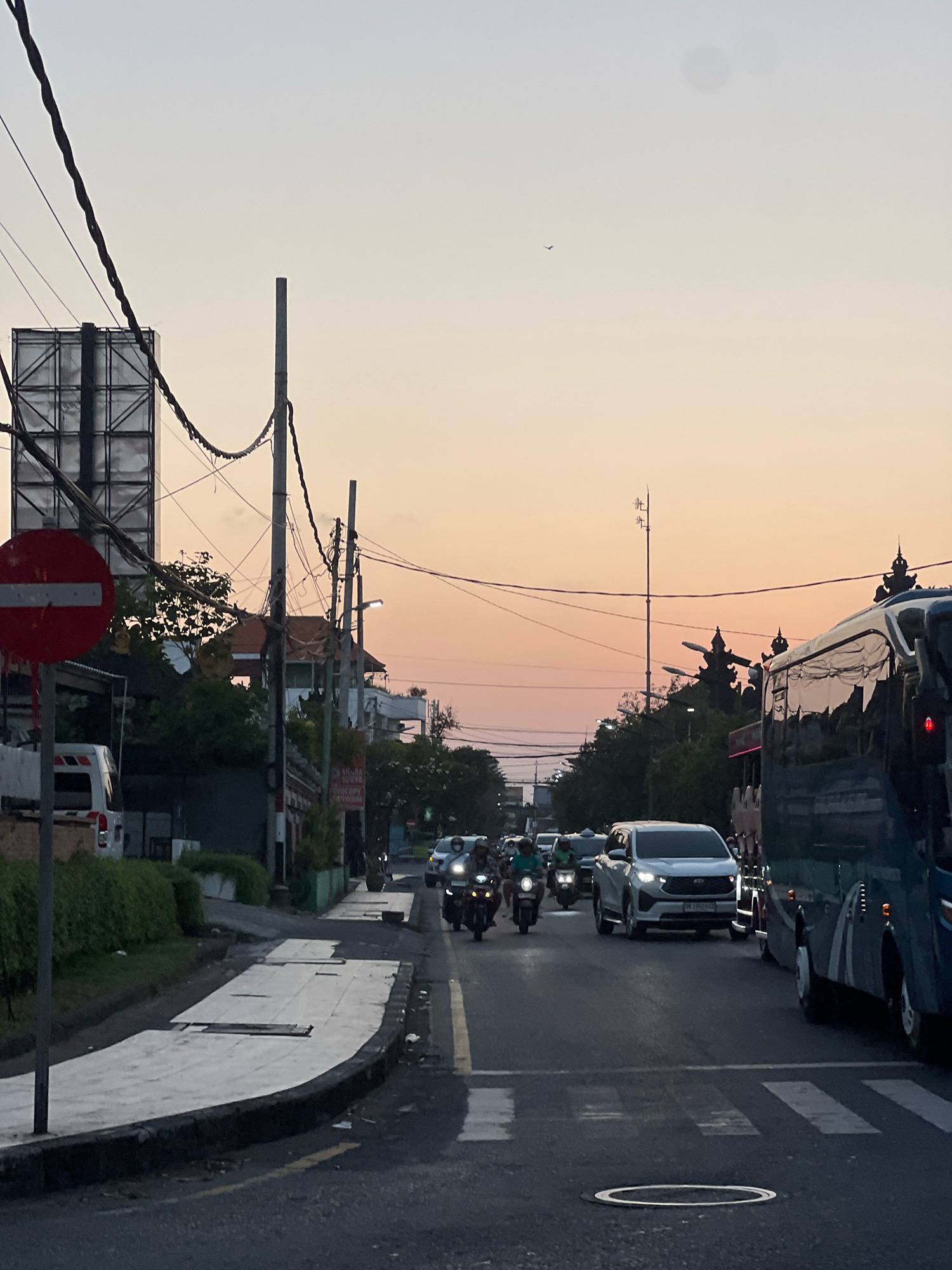 This is a street scene in Kuta, Indonesia, during what appears to be dusk or early dawn. The sky exhibits a gradient of soft orange and pink hues. A busy street is depicted with various vehicles, including motorcycles, cars, and a large bus on the right side of the frame. Several people are visible riding motorcycles. The road surface is asphalt with yellow lane markings and a pedestrian crossing. On the left side of the street, there is a sidewalk with a large circular red and white traffic sign. Buildings and trees line the street. Utility poles with numerous wires are prominent in the foreground and midground, contributing to the urban setting. The overall impression is one of active city life during transitional light conditions.
