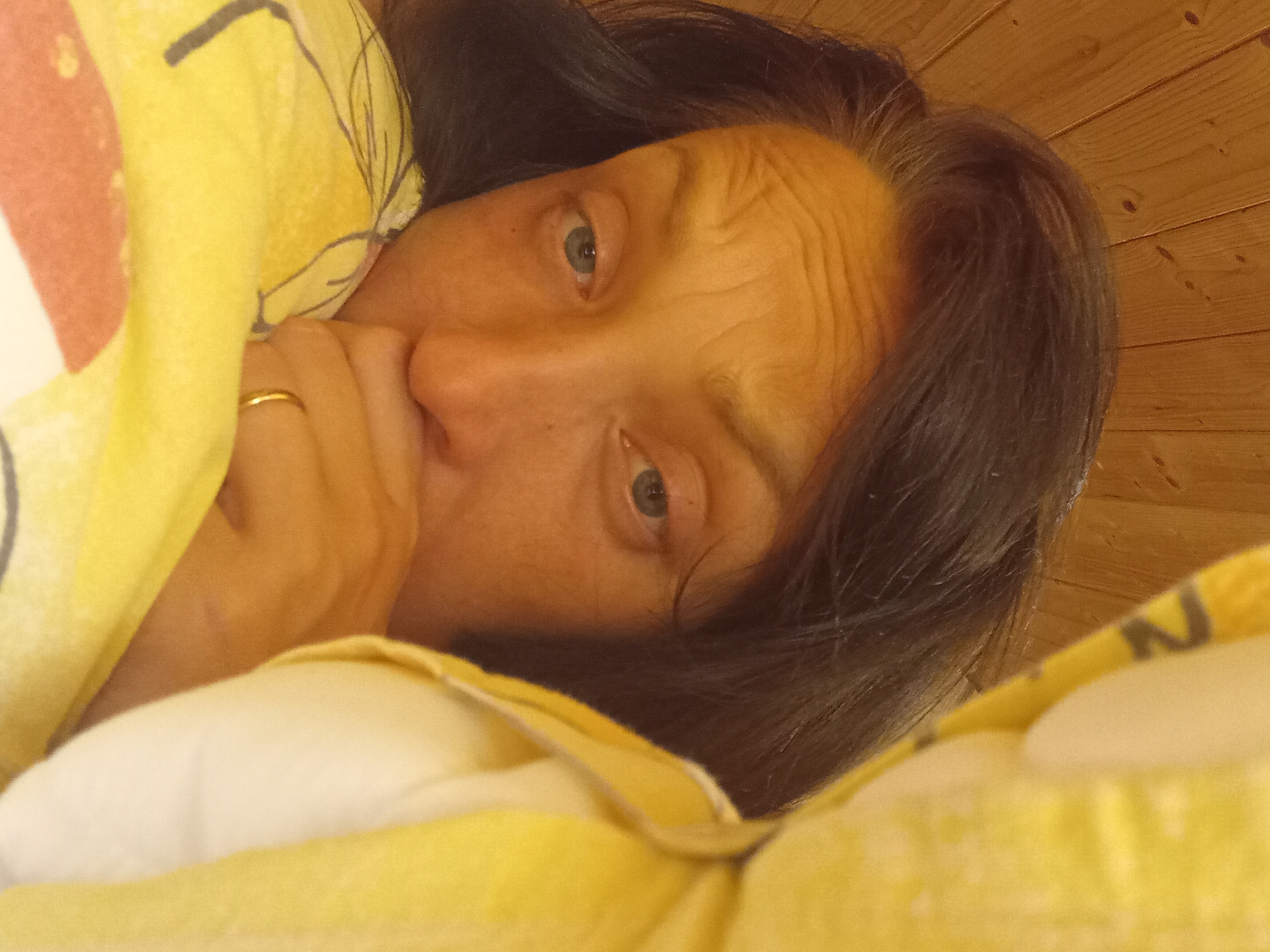 A woman is lying in bed, looking at the camera. She is in a room with wooden walls. She has dark hair and blue eyes. She is wearing a ring on her finger. The bed is covered in a yellow blanket with a pattern of flowers and leaves. The image is a close-up of her face, with her head tilted to the side. The background is blurred, but it appears to be a wooden wall. The overall impression is one of intimacy and vulnerability.