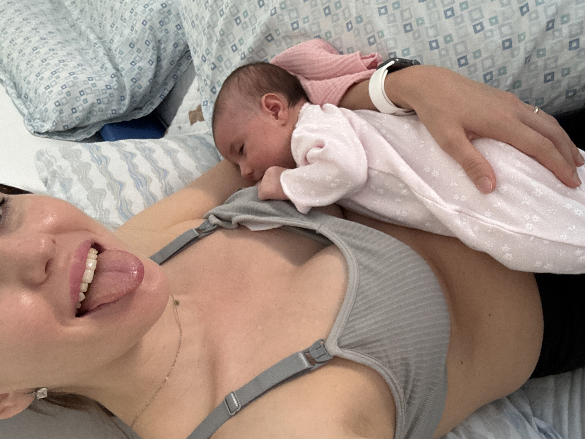 A selfie captures a woman lying down, holding a newborn baby on her chest. The woman is fair-skinned with brown hair, wearing a grey bra-style top and a delicate gold necklace. She is looking at the camera with her tongue out. The baby is dressed in a pink onesie and appears to be sleeping peacefully on her chest. The woman's left hand, adorned with a white smartwatch and a ring, rests gently on the baby. The background reveals a bed with patterned blue and white bedding. The location in Aprilia, Italy is not discernible from the image.