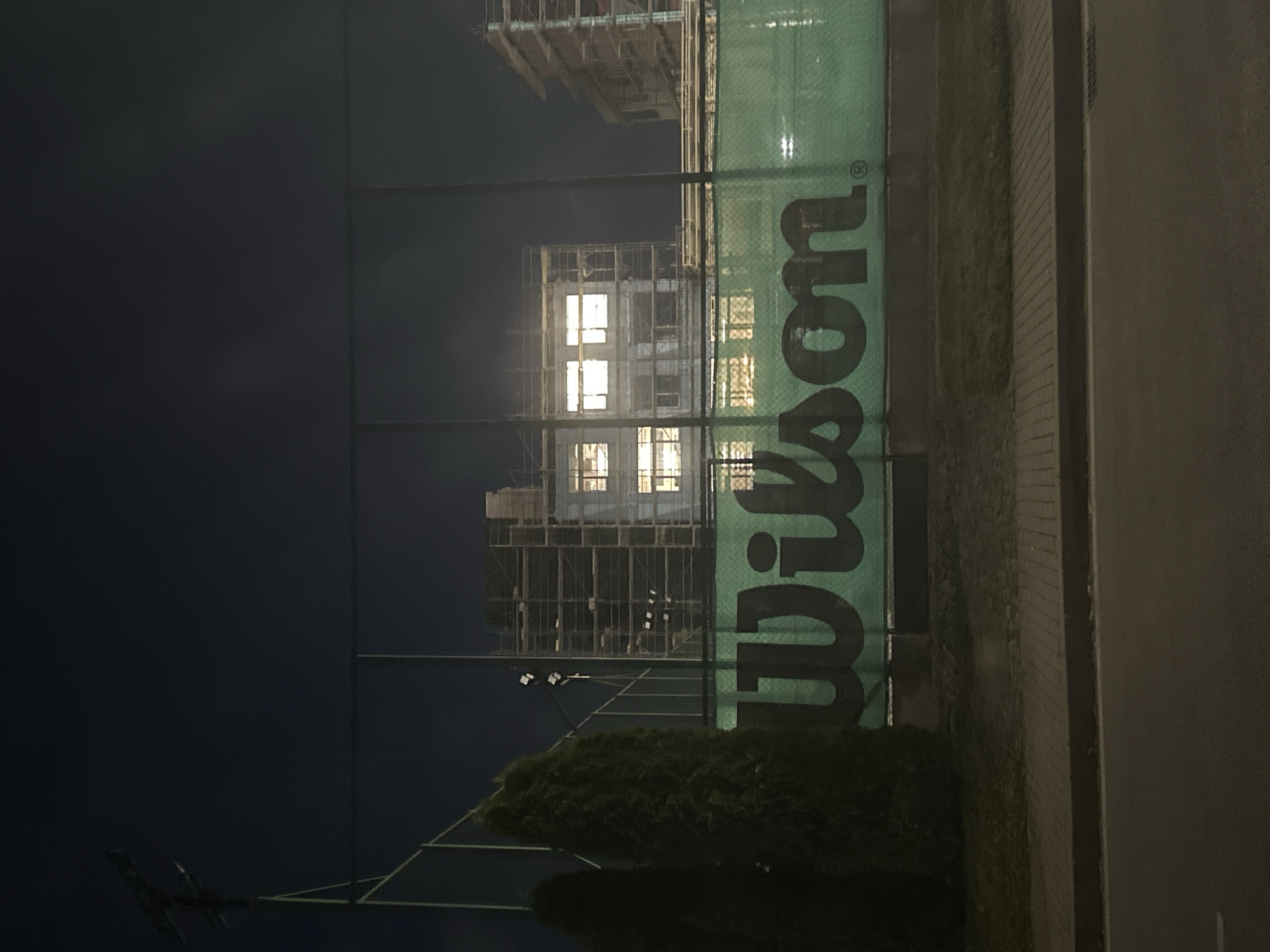The image depicts an outdoor tennis court at night in Sofia, Bulgaria. A green net fence with the "Wilson" logo prominently displayed stretches across the foreground, partially obscuring a construction site in the background. Two dark green evergreen trees stand to the left of the frame. The construction site features buildings covered in scaffolding, with lights illuminating some of the windows. A street and sidewalk are visible in the lower portion of the image. The overall atmosphere is dark and quiet, typical of a night scene in an urban environment.