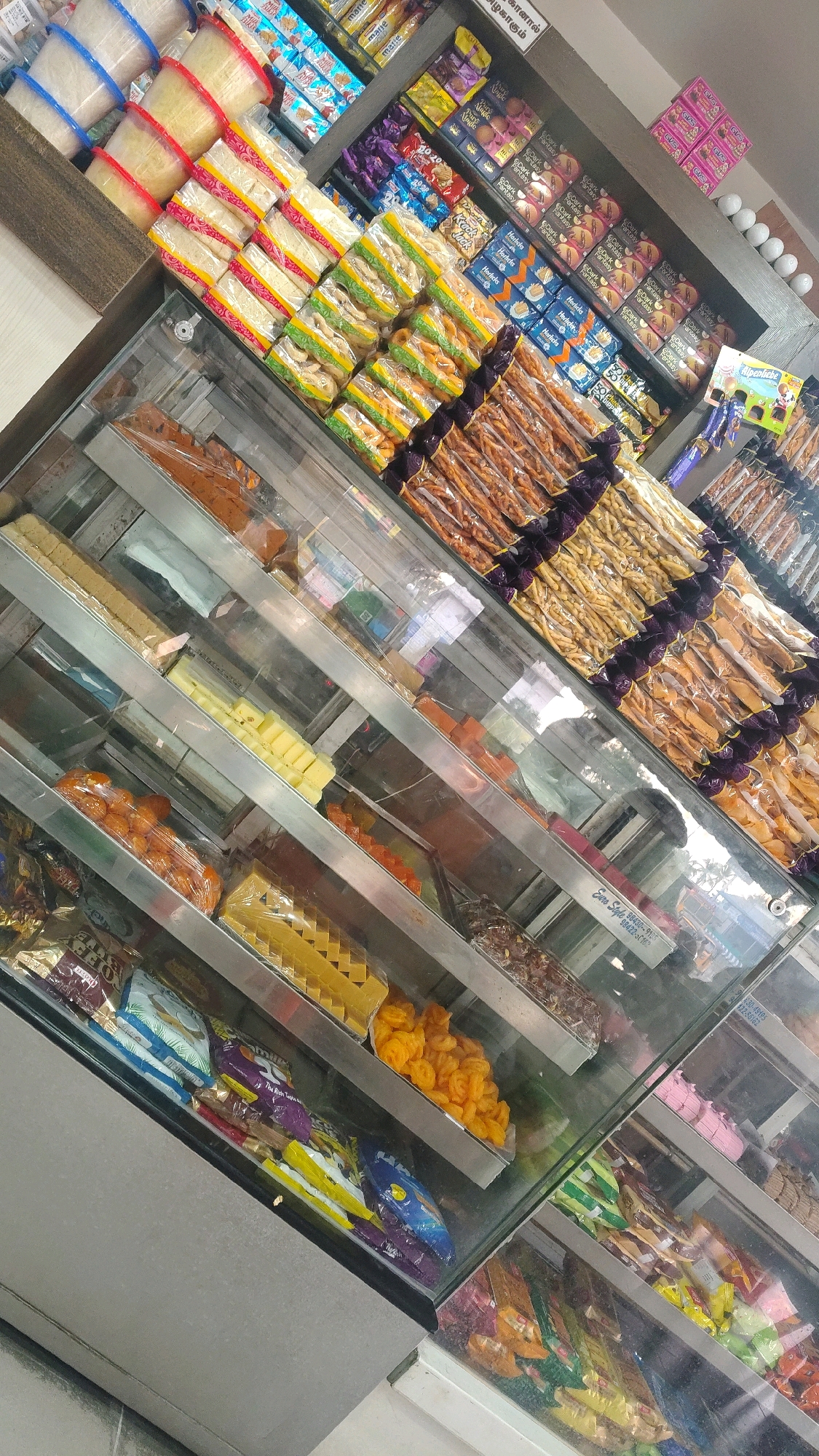 The image shows a display of sweets and snacks in a shop in Dharapuram, India. The display case is filled with various types of confections, including layered sweets, fried snacks, and packaged biscuits. The lighting is bright, and the reflections on the glass case obscure some of the details. The shop appears to be well-stocked and organized.