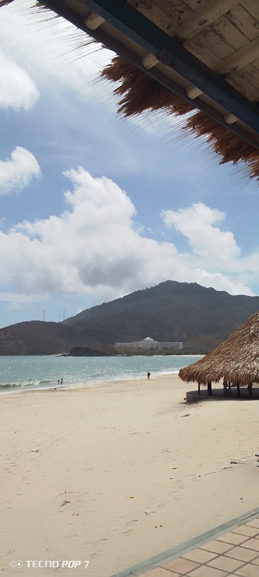 A beach scene in Pedro González, Venezuela, shows a sandy beach, ocean, and mountainous backdrop under a cloudy sky. Two people are in the water, and two others are on the beach. A small thatched-roof structure sits on the beach to the right. In the background, a large white building is visible at the foot of the mountain. A straw-covered structure and part of a tiled surface are in the foreground, partially obscuring the view.