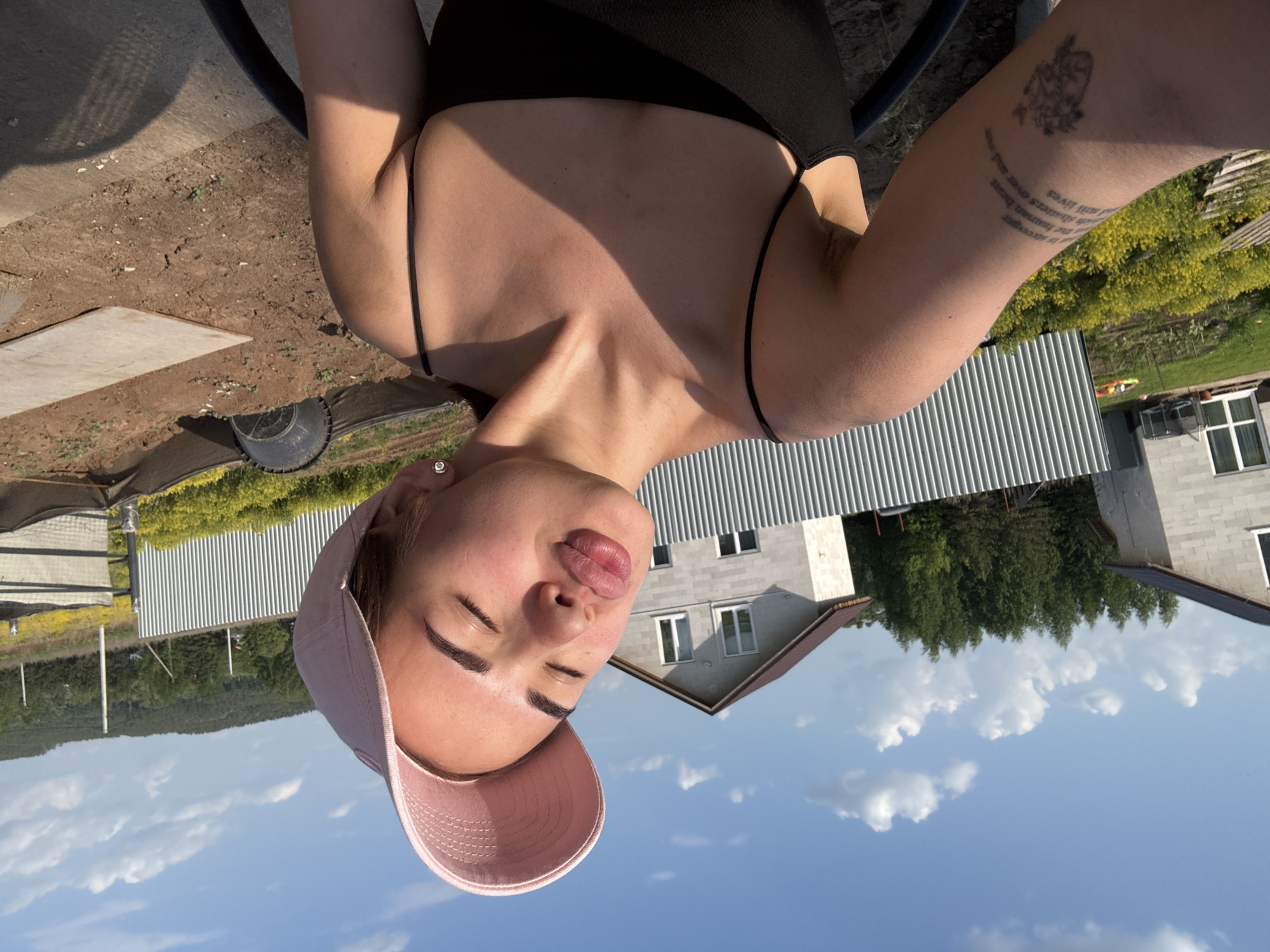 A woman wearing a black top and a pink baseball cap is photographed against a backdrop of a clear blue sky dotted with puffy white clouds. Her eyes are closed, and she has a slight pout. The background includes a two-story light-gray building with a brown roof, surrounded by lush greenery, including trees and a field of yellow flowers. A metal fence is also visible. The scene appears to be in a residential area, resembling the outskirts or a rural part of the city of Лобаново, Russia. The image is oriented in portrait mode.