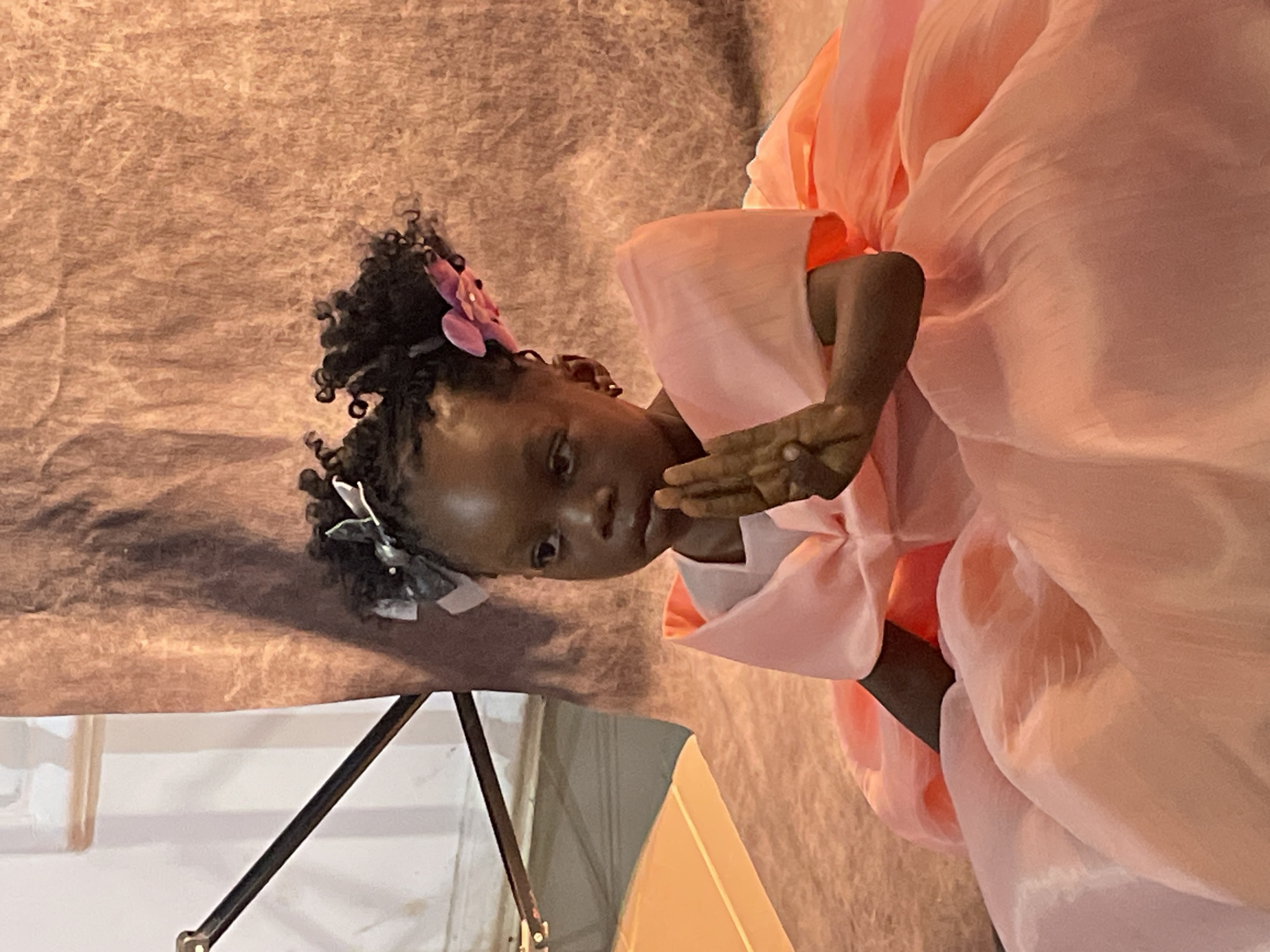 An indoor portrait captures a young girl, likely of African descent, against a textured, light brown or beige backdrop, possibly fabric or a temporary wall. The girl is positioned slightly off-center, her body angled, and her face turned towards the viewer. She wears a striking light pink or peach-colored formal dress with voluminous, flowing fabric, possibly traditional attire like a hanbok given its style. Her dark, curly hair is styled into two pigtails, adorned with a delicate pink flower on one side and a grey and white bow on the other.
The girl has a thoughtful or pensive expression, with her dark eyes looking directly forward. Her right hand is raised, with fingers gently touching her chin or cheek, a gesture that adds to her introspective demeanor. The lighting is soft, highlighting the sheen of her dress and the textures of the backdrop. In the lower left corner, parts of dark metal stands or structures are visible, suggesting an event setting or a stage for a performance or presentation. There is no visible text in the image. The scene takes place in Jos, Nigeria, hinting at a local cultural event or celebration where such attire might be worn.