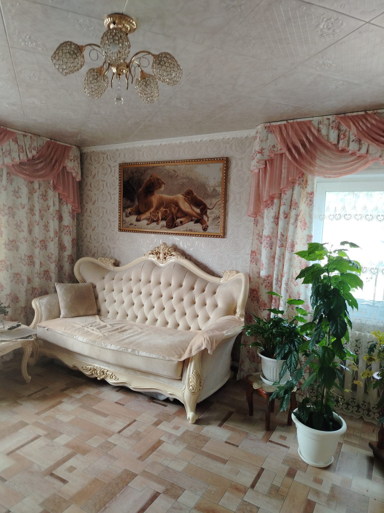 The image shows a living room interior. A plush, cream-colored sofa with tufted upholstery and ornate gold trim is the central piece of furniture. A framed painting of lions rests on the wall above the sofa. To the right of the sofa, there are several potted green plants, with the largest one being tall and leafy. Sheer pink and floral patterned curtains adorn the window. A decorative chandelier with crystal-like elements hangs from the ceiling, which has a patterned texture. The floor is covered in a light-colored tile with a geometric pattern. The lighting suggests daytime. No people are visible in the image. This scene is located in Rusalevka, Russia.