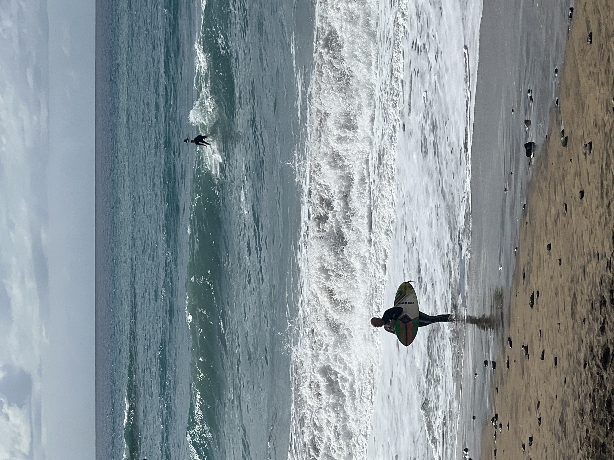 This image captures a surfing scene on a beach in Las Palmas, Spain, under an overcast sky. The horizon line stretches across the mid-left, separating the light grey, cloudy sky from the vast expanse of the ocean.
Two surfers are visible in the frame. Further out in the ocean, a surfer in a dark wetsuit is paddling on a surfboard, appearing to ride or approach a small wave. Closer to the foreground, on the right side, another surfer in a dark wetsuit stands on the wet sand at the water's edge. This surfer holds a distinctive surfboard vertically. The board features vibrant green, red, and black striped patterns, and prominently displays the white text "LSD" on its underside. The sandy beach is scattered with numerous dark pebbles and rocks, especially near the waterline, where the white foam of breaking waves meets the shore. The ocean water transitions from a deep blue further out to frothy white as waves crash towards the beach. The scene depicts typical daytime surfing conditions.