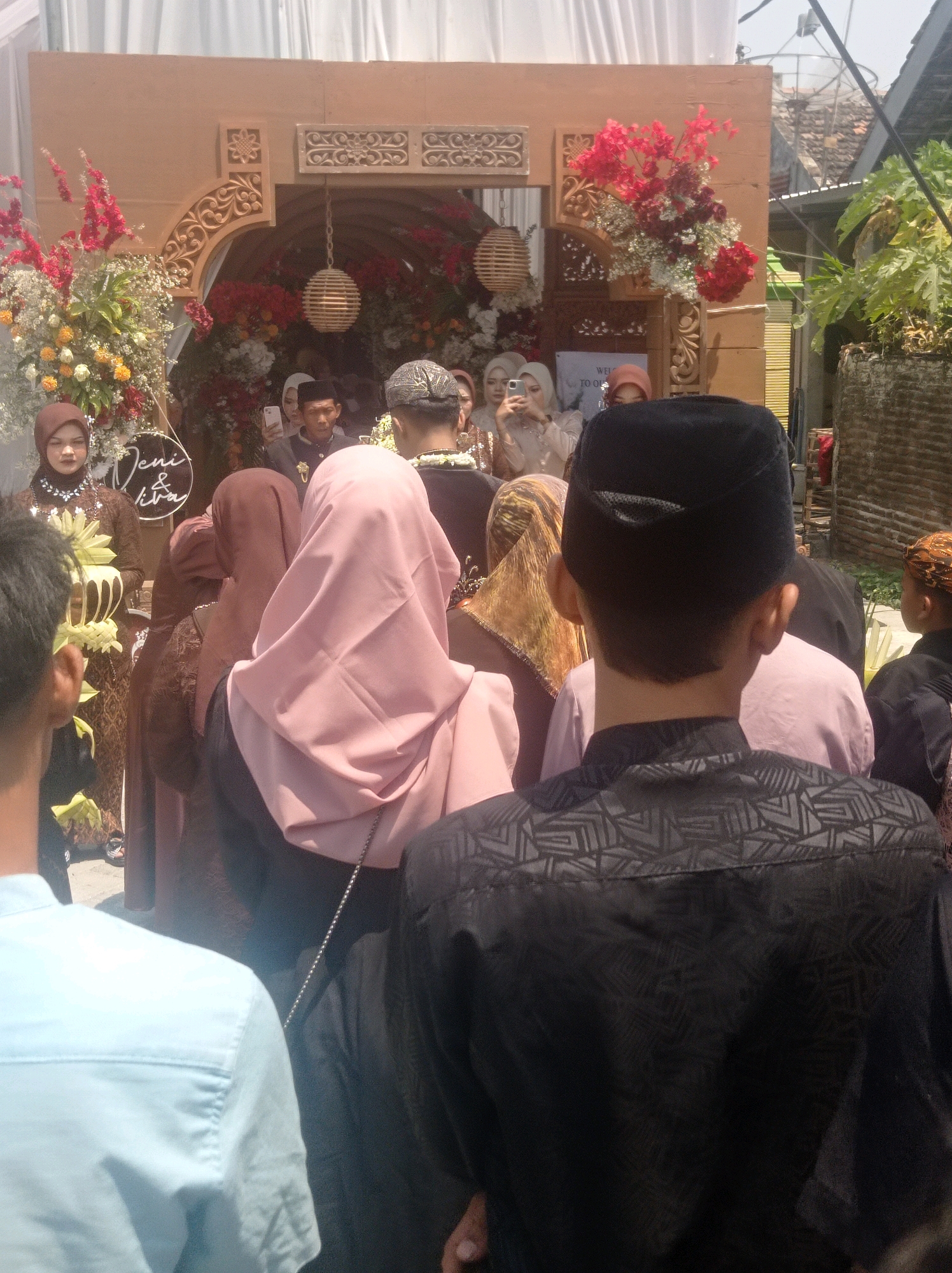 A wedding ceremony is taking place outdoors in Ngemplak, Indonesia. The scene features a decorated archway with red and white flowers. Several individuals are present, some wearing traditional attire. A woman in a brown hijab is holding a bouquet. Another woman in a pink hijab is also visible. In the foreground, a person wearing a black patterned shirt faces away from the camera, and another person in a light blue shirt is also seen from the back. The background shows a brick wall and lush green foliage, suggesting a residential or garden setting. A white banner with text is affixed to the archway.