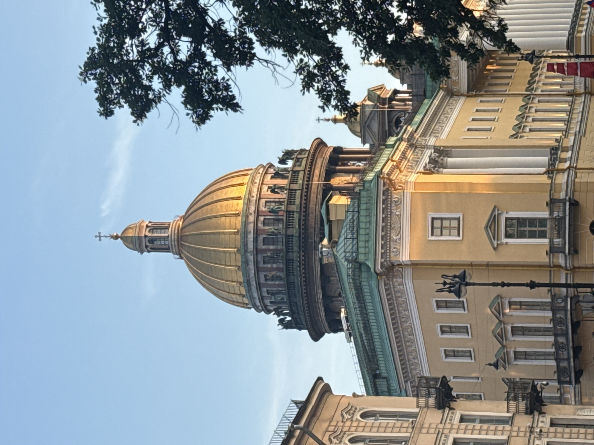 The image shows a view of Saint Isaac's Cathedral in Saint Petersburg, Russia, under a clear blue sky. The cathedral's prominent golden dome is illuminated by the sun, casting a warm glow on its ornate architecture. The lower sections of the cathedral, with their green roofs and yellow facades, are also visible, along with surrounding buildings that feature balconies and classical architectural elements. A large, dark green tree in the foreground partially obscures the view of the cathedral and the sky. The scene appears to be captured during daylight, likely in the late afternoon or early morning, given the angle of the sunlight. No people are visible in the image.