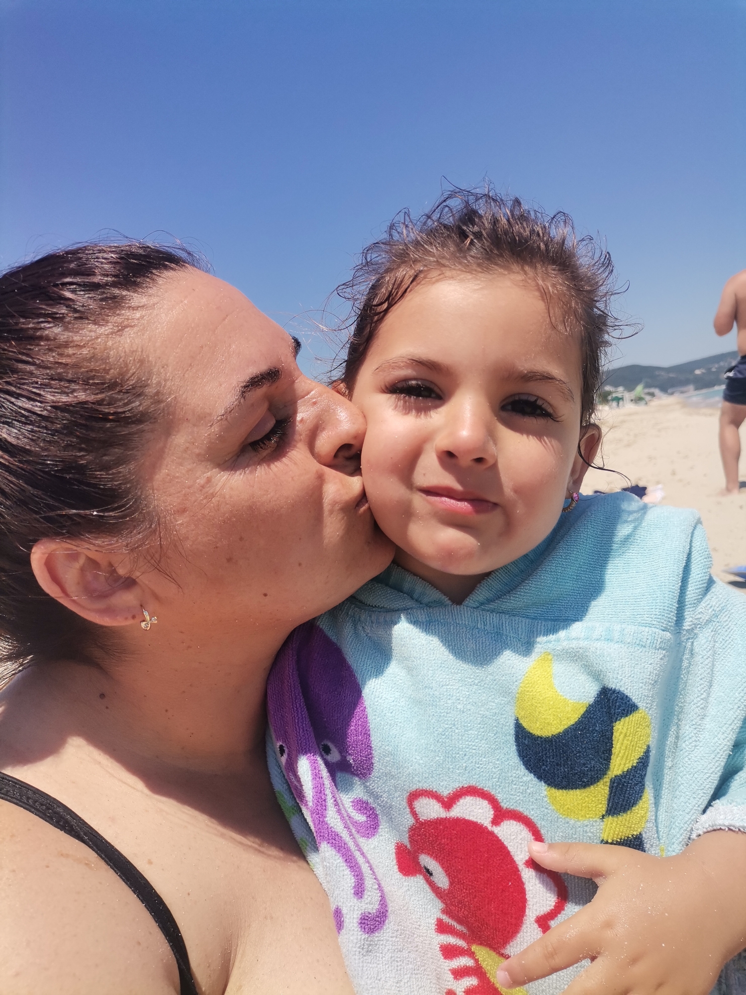 This media file shows a close-up of a woman kissing a young girl on the cheek. They are at a beach, with the sea and a sandy shore visible in the background, under a clear blue sky. The weather appears to be sunny and warm, suggesting it's daytime.
The woman, on the left, has dark hair and is wearing a black swimsuit. She has a small hoop earring in her left ear. The girl, on the right, is wearing a light blue hooded towel with a cartoon bee and fish design. Her hair is dark and curly, and she has a gentle smile. The girl's face is directed towards the camera, while the woman's face is turned towards the girl, capturing a tender moment between them.
The background shows a sandy beach with some beach umbrellas and people in the distance, implying a public beach. The sea is calm and the sky is bright blue, indicating a pleasant summer day. The overall mood is affectionate and joyful, highlighted by the intimate gesture and the bright, sunny setting. The location context provided is Kranevo, Bulgaria. There is no visible text in the image.
