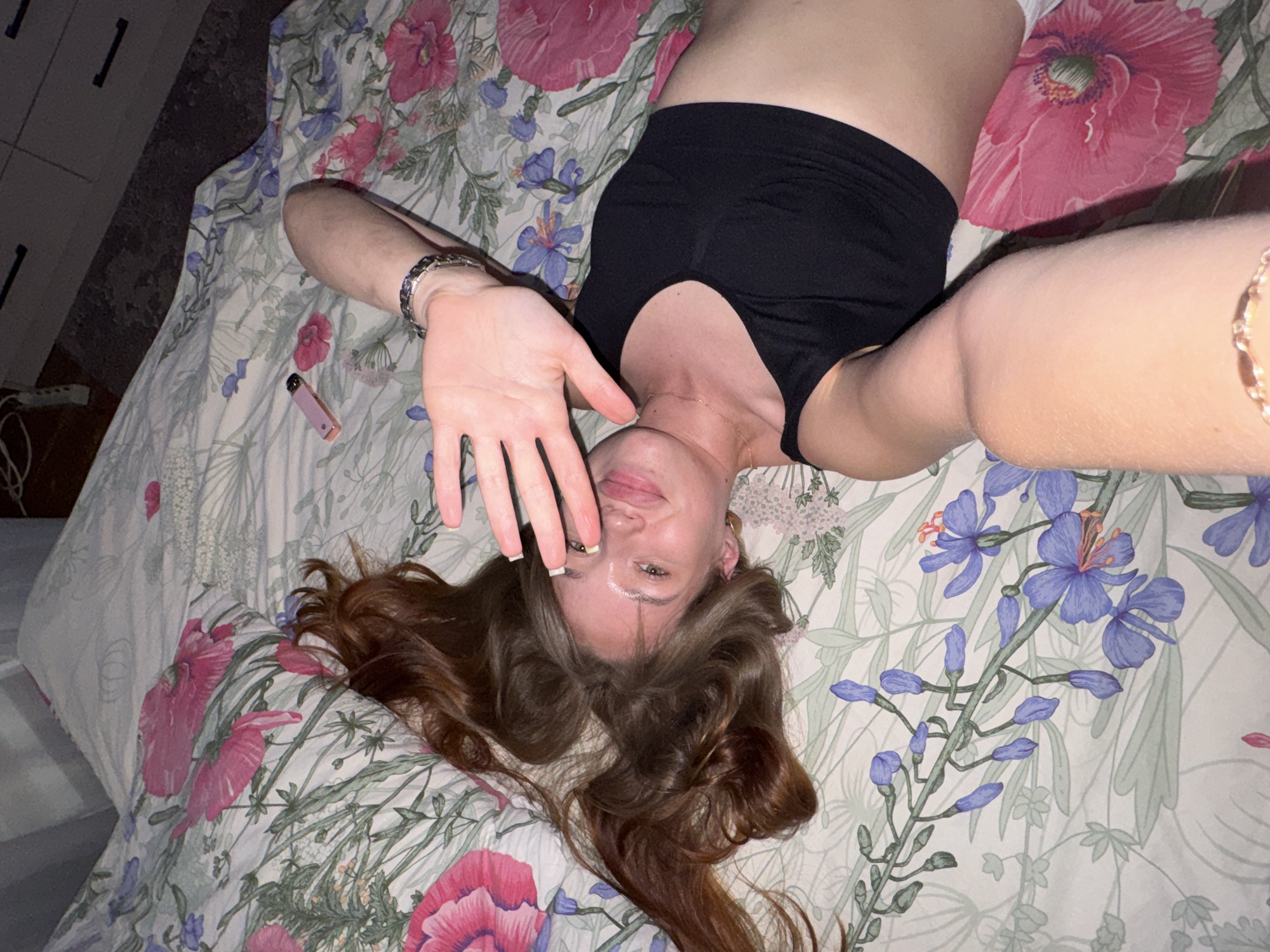 A young woman with long, reddish-brown hair lies on her back on a bed with floral-patterned bedding. She is wearing a black sports bra. Her right hand is raised to partially cover her face, and she is looking towards the camera with a slight smile. Her left arm is bent at the elbow, resting on her stomach. A pink vaping device is visible on the bedding near her head. The bedding features large pink flowers and smaller blue flowers interspersed with green foliage. The scene appears to be indoors, likely in a bedroom. The image is a selfie, taken from a slightly elevated angle looking down at the woman. There are no specific landmarks visible that would indicate the city of Chaikovsky, Russia.